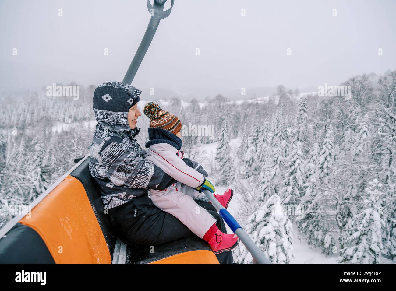 Mutter mit einem kleinen Kind auf dem Schoß in Skianzügen geht mit einem Sessellift den Berg hinauf Stockfoto