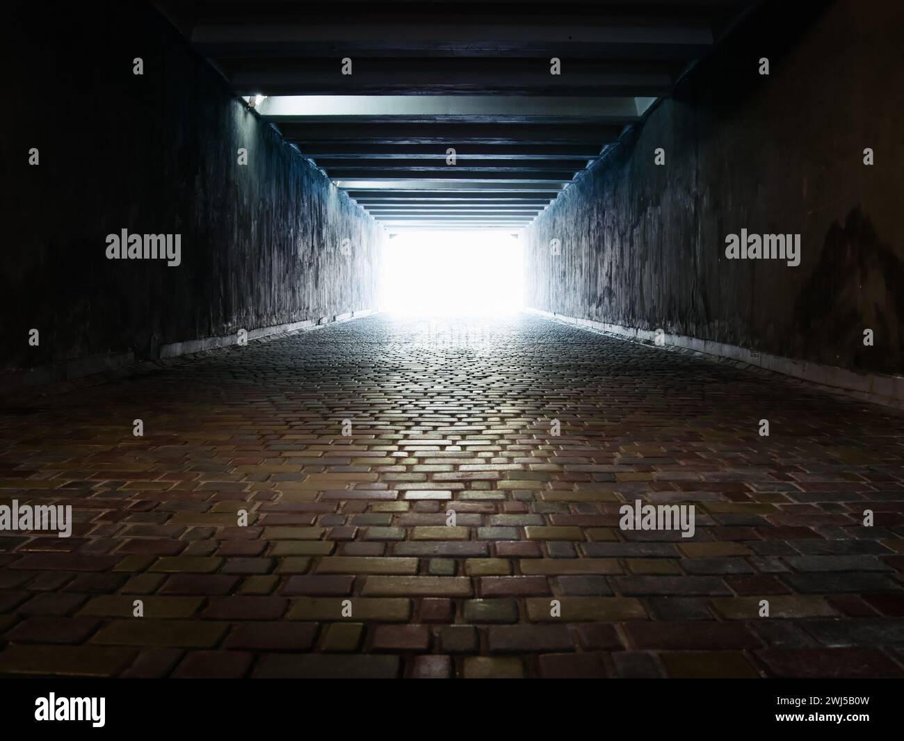 Licht am Ende eines dunklen Fußgängertunnels, der mit mehrfarbigen Pflastersteinen gepflastert ist. Hintergrund Stockfoto