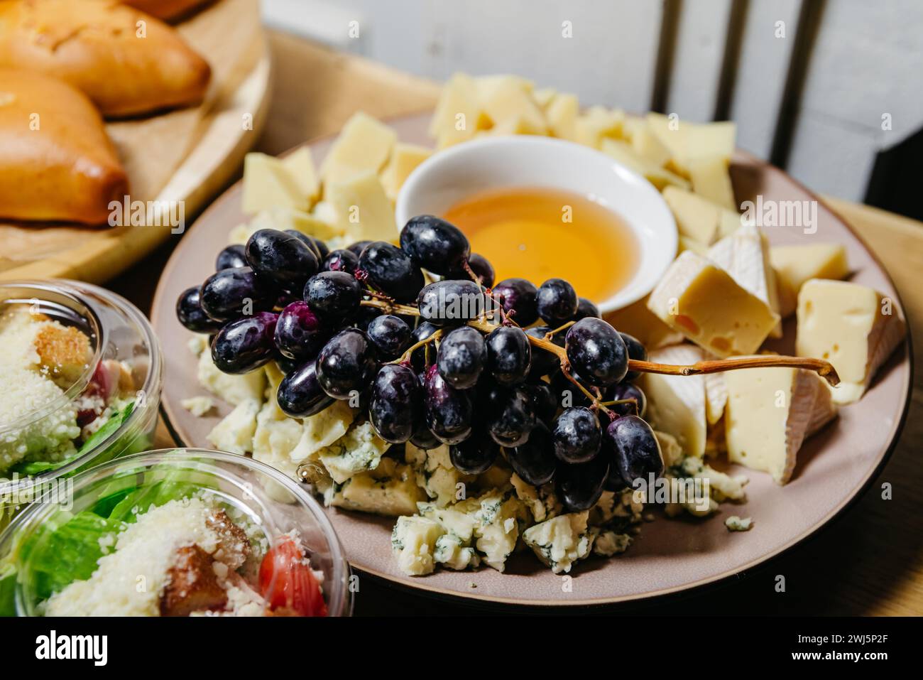 Eine Auswahl an Gourmet-Käsesorten, begleitet von frischen schwarzen Trauben und einer Schüssel Honig, perfekt für eine Käseplatte oder eine Weinpaarung. Stockfoto