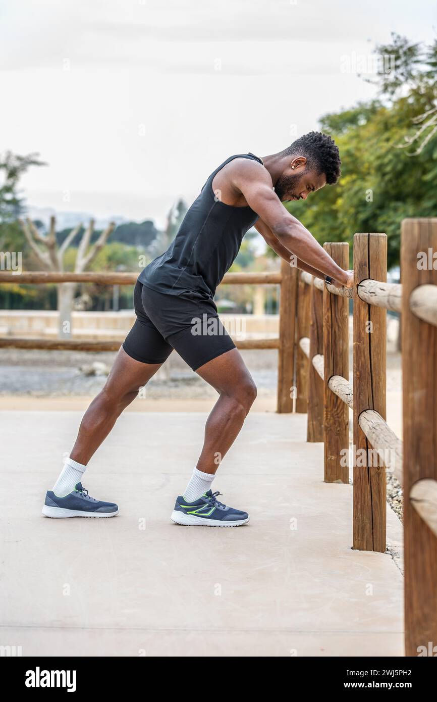 Ganzkörper-Seitenansicht eines jungen afroamerikanischen männlichen Athleten in Aktivkleidung, der die Beine beim Aufwärmen vor dem Training dehnt Stockfoto