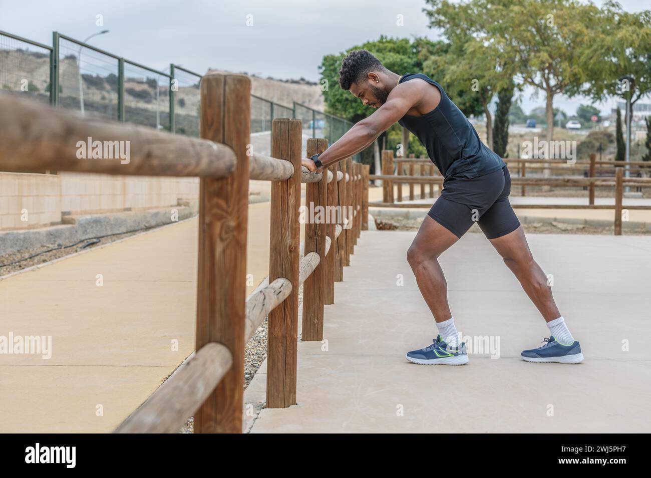 Seitenansicht Ganzkörper eines starken afroamerikanischen Sportlers in schwarzer Sportbekleidung, der sich auf einen Holzzaun stützt und sich vor dem Fitnesstraining aufwärmt Stockfoto