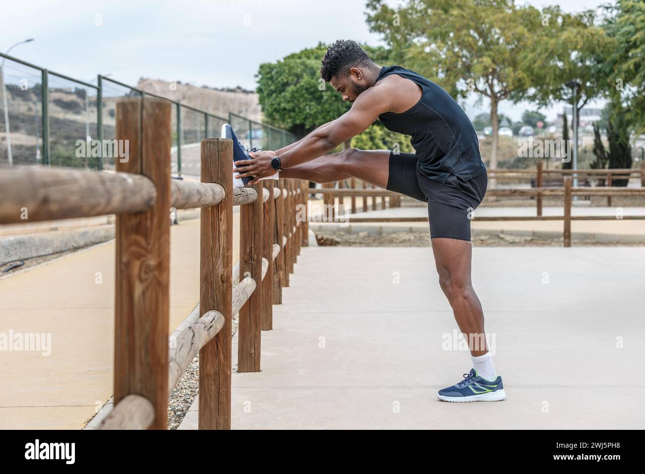 Ganzkörper-Seitenansicht eines starken afroamerikanischen Athleten, der das Bein auf einen Holzzaun legt und während des Fitnesstrainings mit den Zehen trainiert Stockfoto