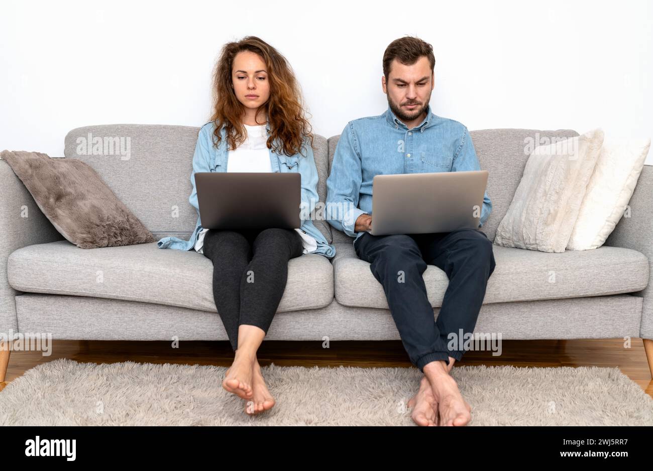 Seriöse und fokussierte Mann und Frau sitzen nebeneinander auf dem Sofa und benutzen Laptops. Stockfoto