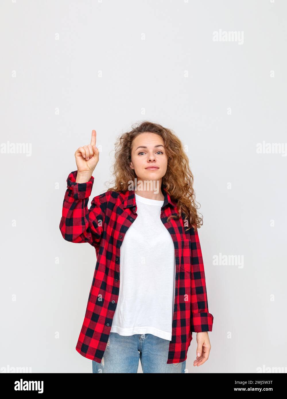 Lockige brünette Frau in lässigem Outfit, die vor dem Hintergrund im Studio steht und mit dem Finger nach oben zeigt. Stockfoto