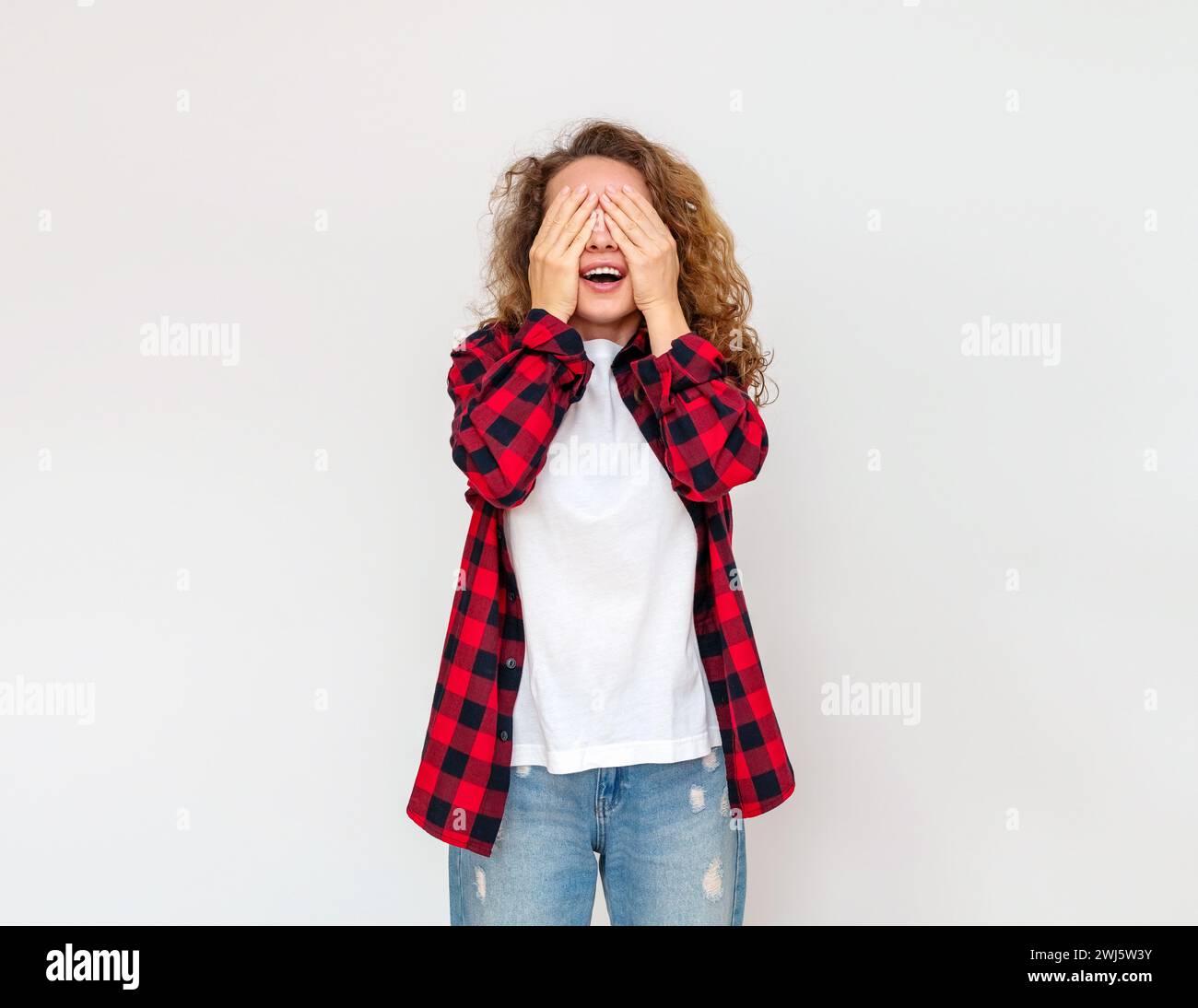 Lockige Frau in lässiger Kleidung, die im Studio steht und die Augen mit den Händen bedeckt. Stockfoto