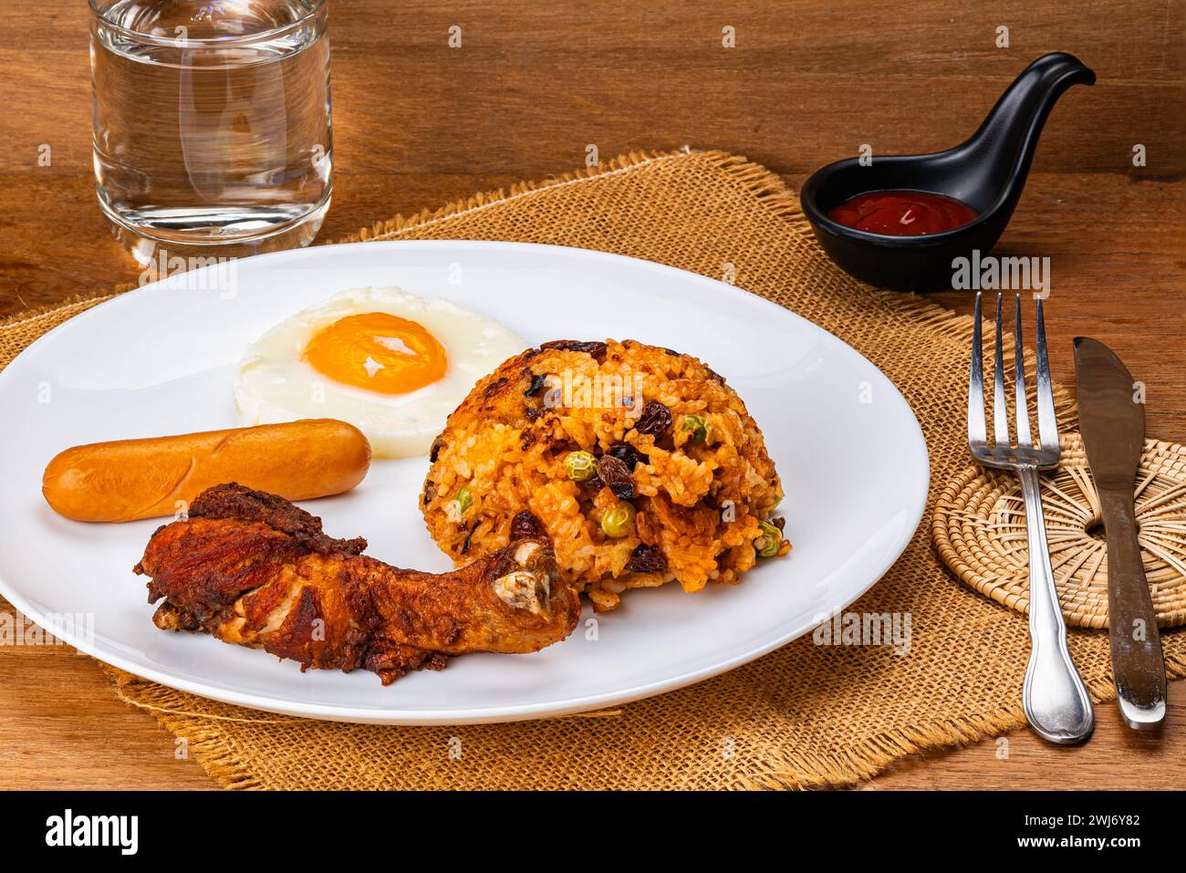Amerikanischer gebratener Reis mit gebratenem Trommelstock, Spiegelei und Wurst auf weißer Keramikplatte mit Ketchup in schwarzer Tasse, einem Glas Wter, Metallgabel und Messer Stockfoto