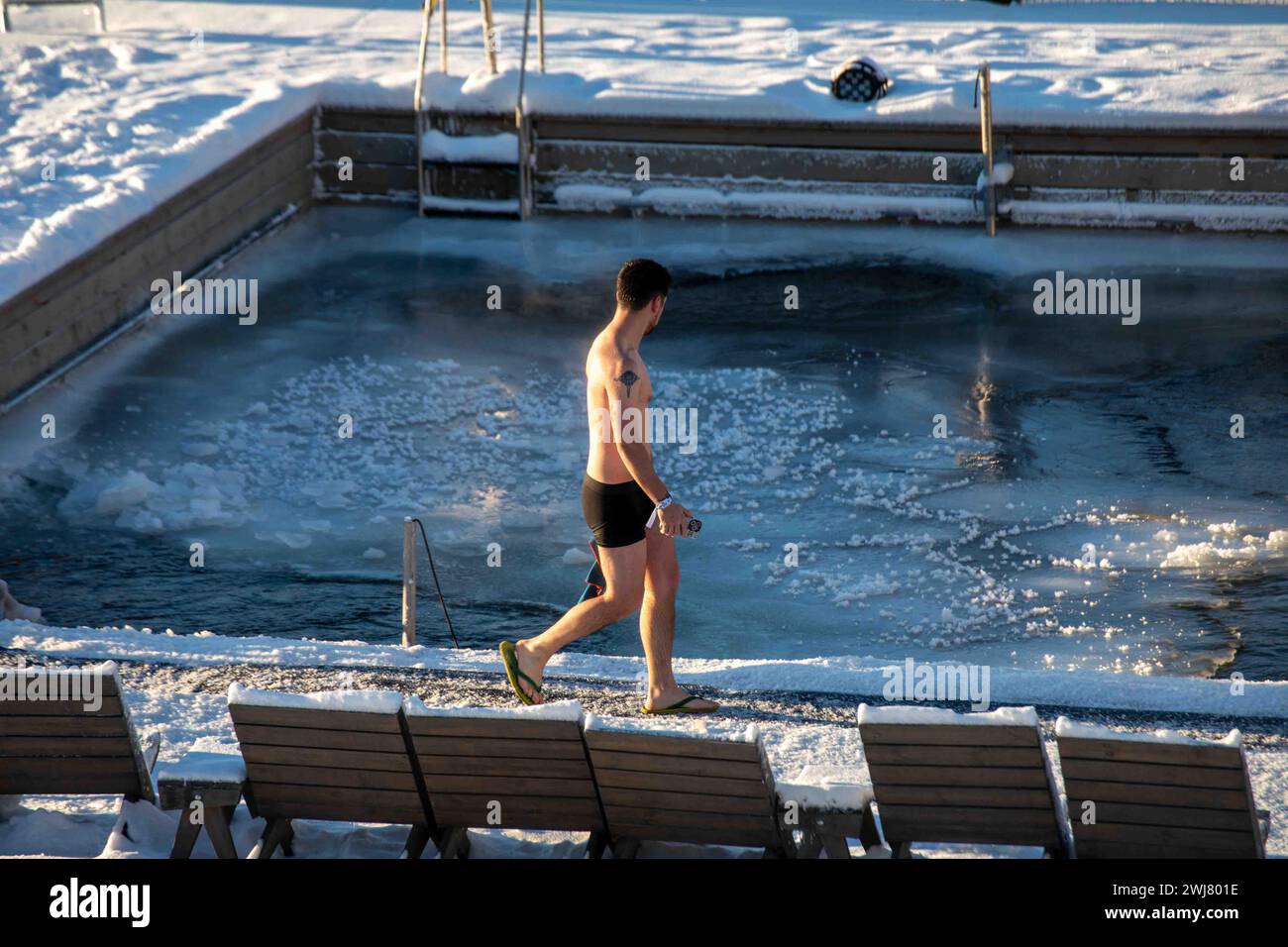 Mann, der an einem sonnigen Wintertag im Stadtteil Katajanokka in Helsinki, Finnland, vorbei an einem gefrorenen Meerwasserpool im Allas Sea Pool, einem Meeresbad und lido, spaziert Stockfoto