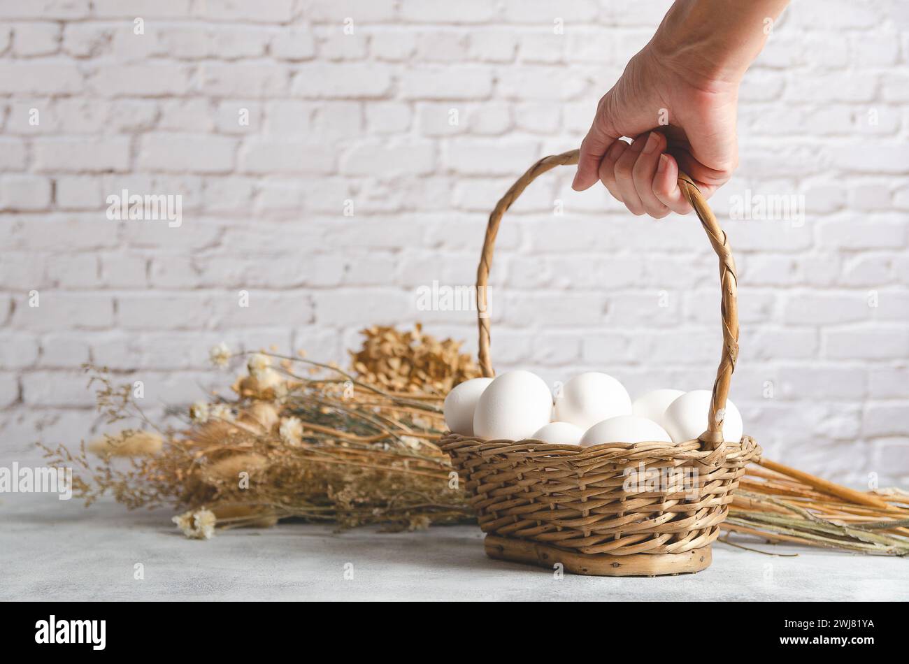Weibliche Hand, die einen Korb mit weißen Hühnereiern auf einem hellen Hintergrund mit getrockneten Blumen hält. Vorderansicht mit Kopierraum. Stockfoto