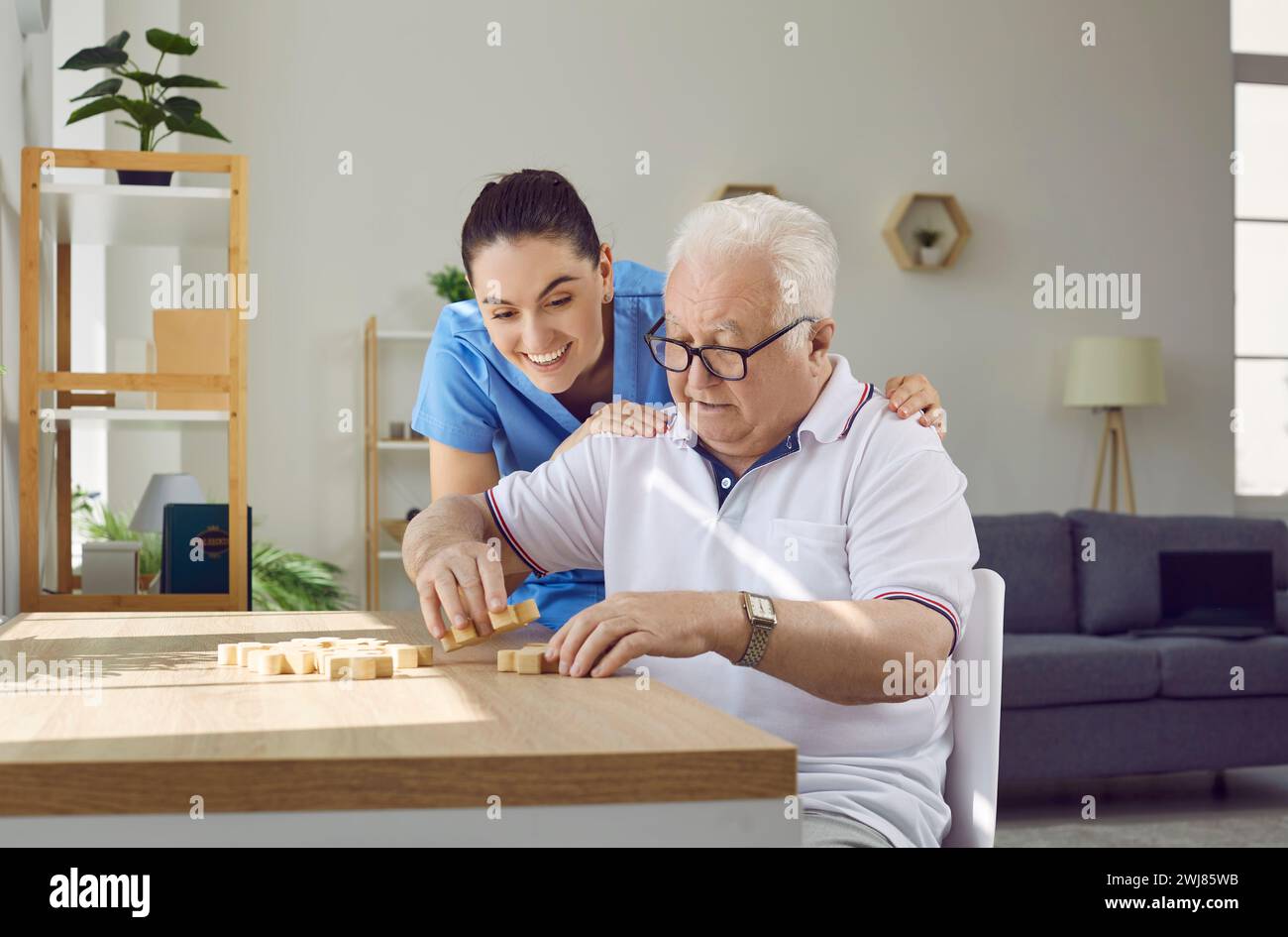 Freundliche, fürsorgliche Krankenschwester hilft älteren Männern, Holzpuzzleteile richtig zu kombinieren und zu montieren. Stockfoto