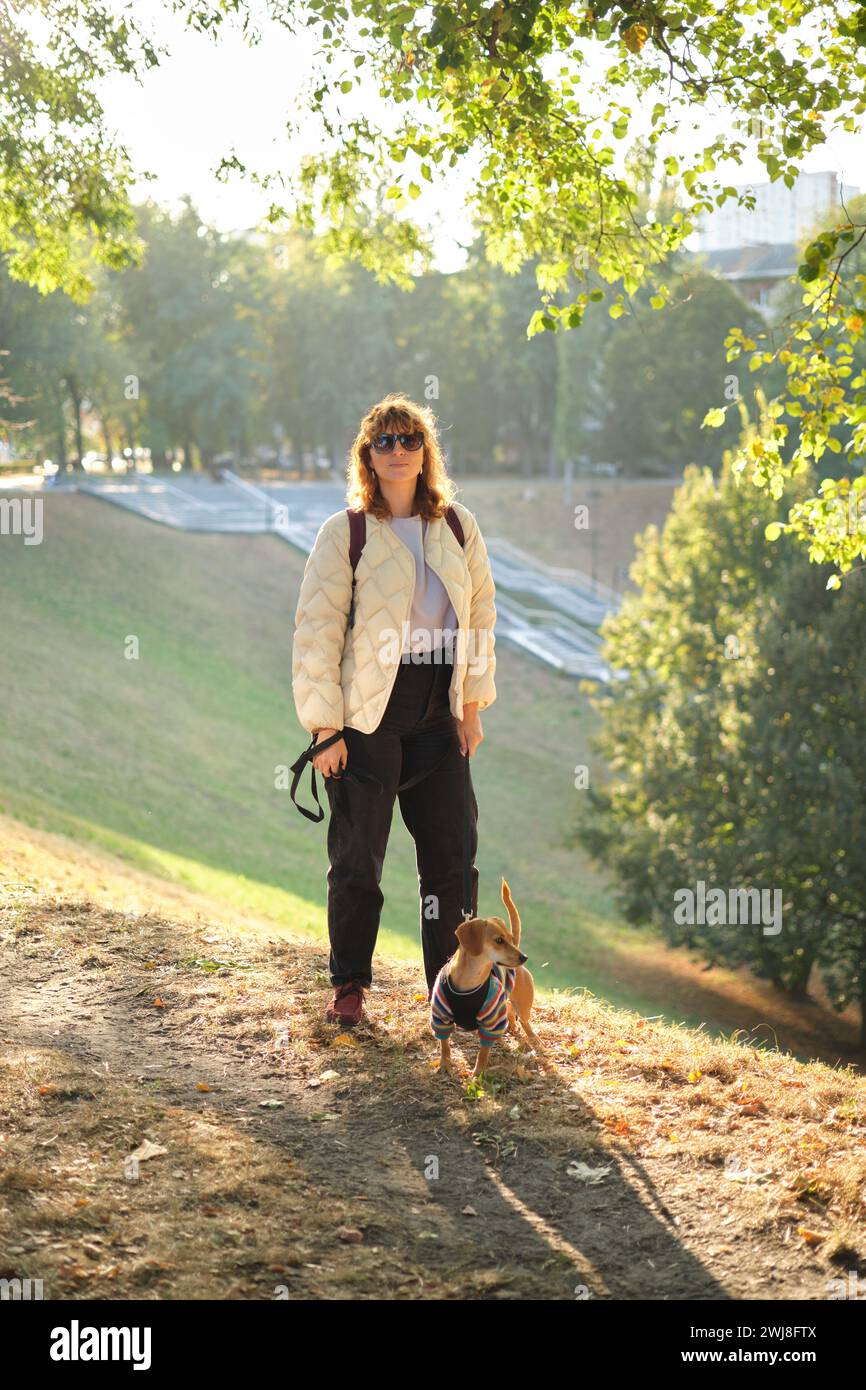 Kaukasisches Mädchen Dackelbesitzerin, die mit Hund im Park draußen läuft, Herbstferien. Haustierkonzept. Freundschaft zwischen Mensch und Haustier. Lustiger Welpe mit Pullover. Hochwertiges vertikales Foto Stockfoto