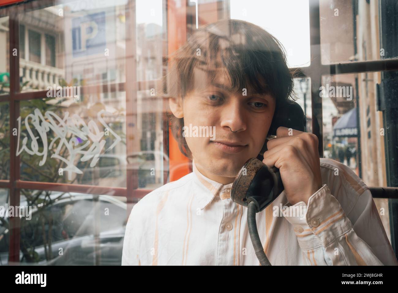 Junger stilvoller Mann im Stil der 90er Jahre, der in rotem Retro-altmodischen Telefonkabine in der Stadtstraße mit Reflexion im Telefonkastenglas spricht Stockfoto