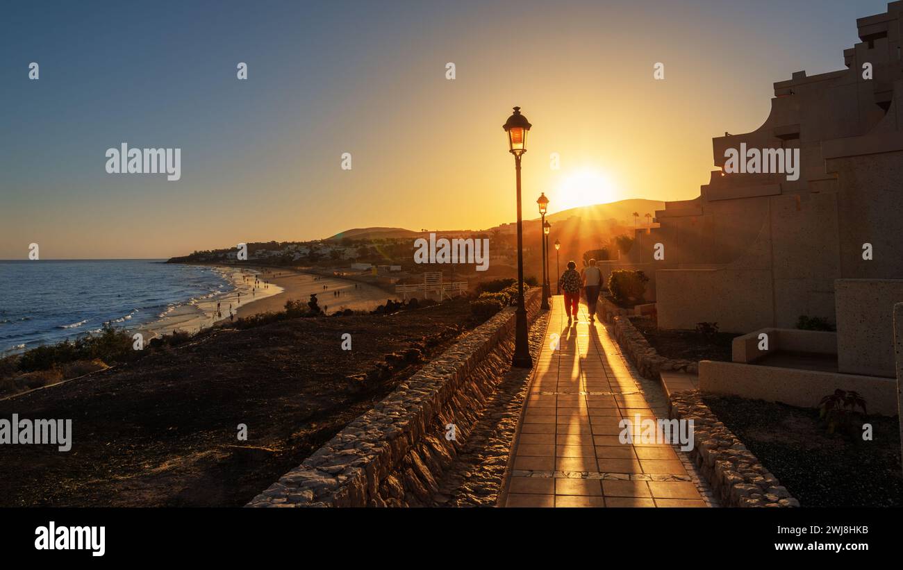 Ein paar Touristen gehen entlang eines Spaziergangs, der bei Sonnenuntergang von strahlendem Sonnenlicht beleuchtet wird, am Strand Costa Calma auf der Insel Lanzarote auf den Kanarischen Inseln Stockfoto