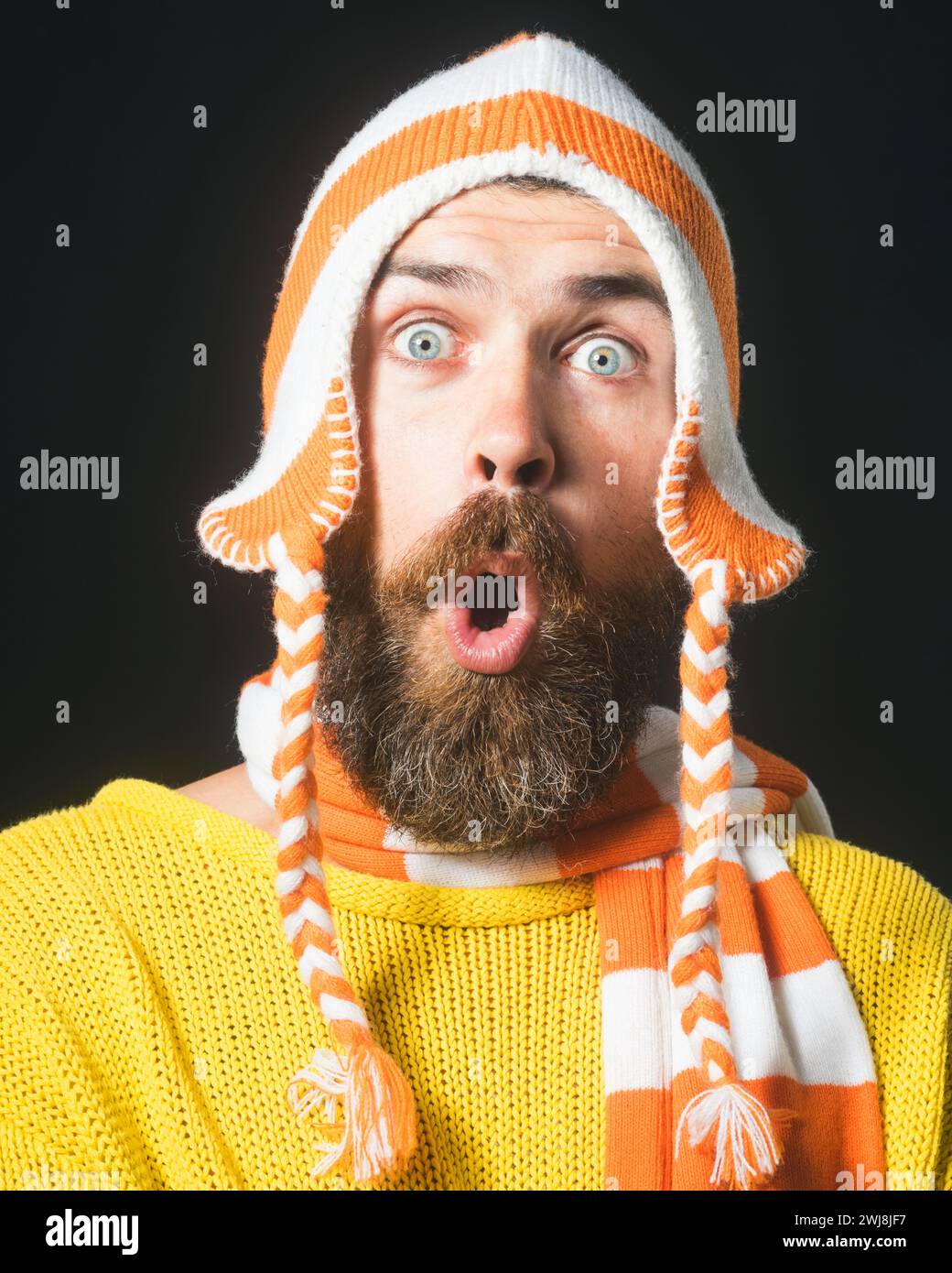 Porträt eines überraschten bärtigen Mannes in gelbem Pullover, gestreiftem lustigem Hut und Schal. Winteraccessoires. Ein schöner, staunender Mann mit Bart und Schnurrbart Stockfoto