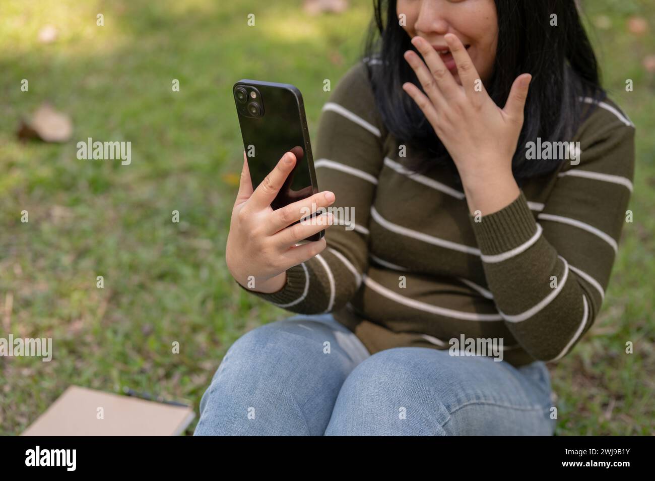 Eine Nahaufnahme einer aufgeregten asiatischen Frau, die eine unerwartete Nachricht auf ihrem Handy liest, während sie sich in einem Park entspannt. Stockfoto