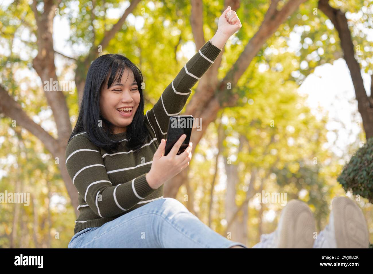 Eine fröhliche junge asiatische Frau schaut mit einem aufgeregten, glücklichen Gesicht auf ihr Smartphone und hebt als Reaktion auf gute Nachrichten die Hand, während sie auf einem Be sitzt Stockfoto