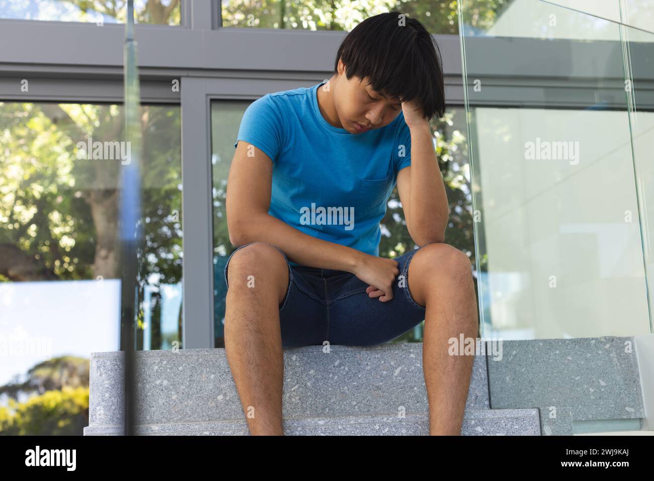 Der Teenager asiatische Junge sitzt vor einem modernen Gebäude und sieht nachdenklich und traurig aus Stockfoto