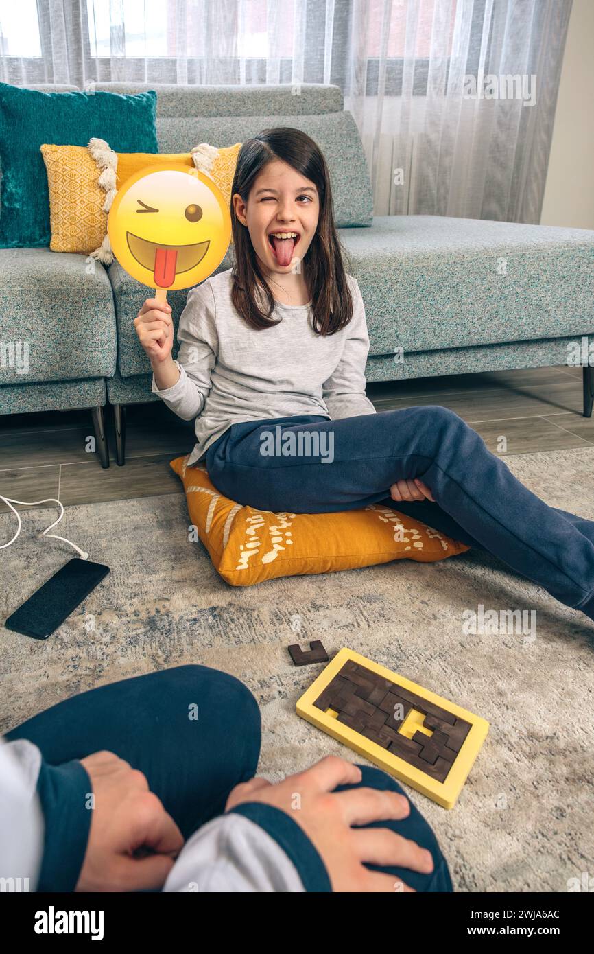 Ein junges Mädchen streckt verspielt ihre Zunge heraus, während es ein blinkendes Emoji-Gesichtsschild hochhält und auf einem Kissen in einem lebendigen Wohnzimmer sitzt Stockfoto