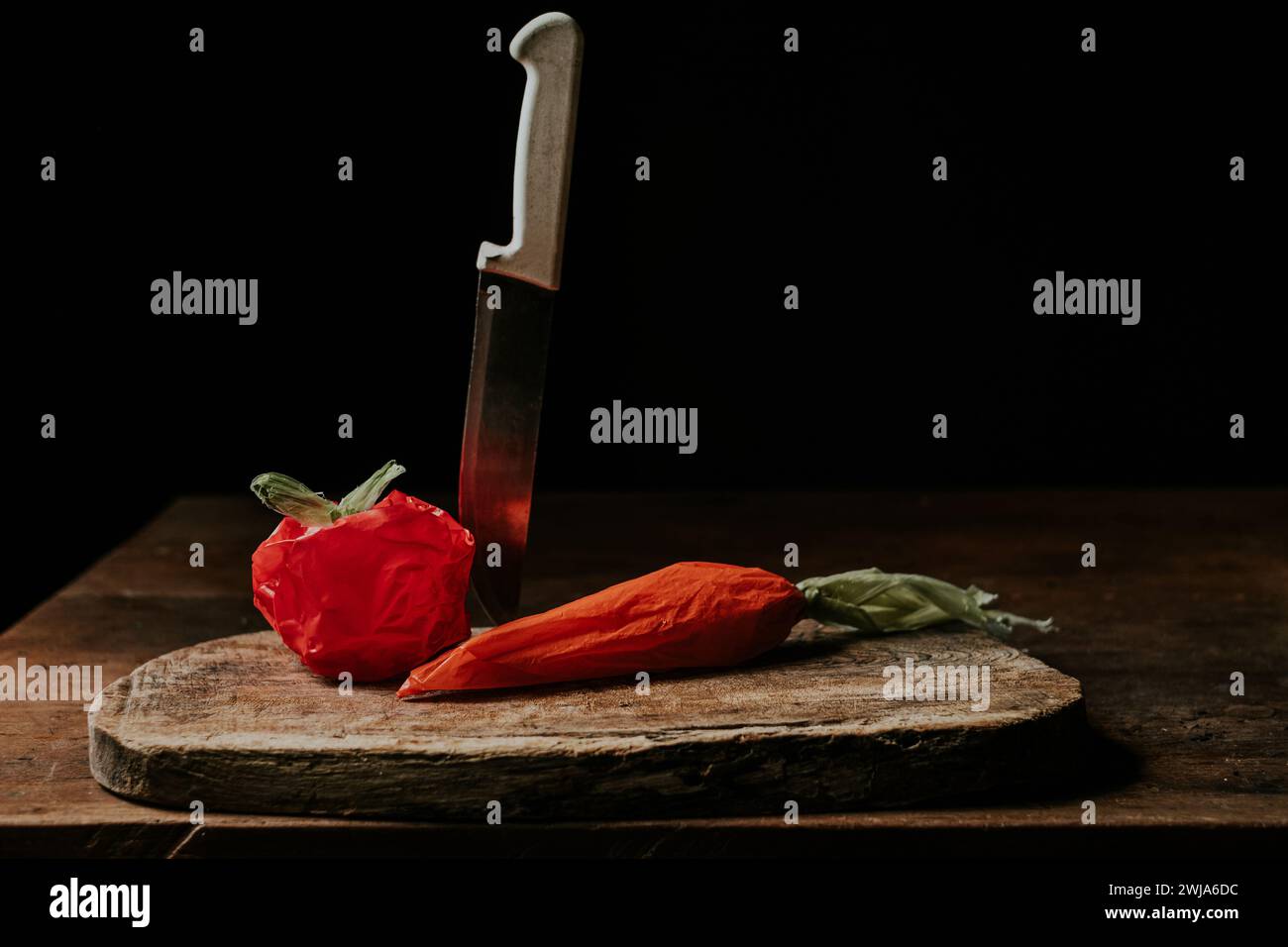 Plastiktüten, die an eine Tomate und Karotte erinnern, neben einem Küchenmesser auf einem rustikalen Holzschneidebrett Stockfoto