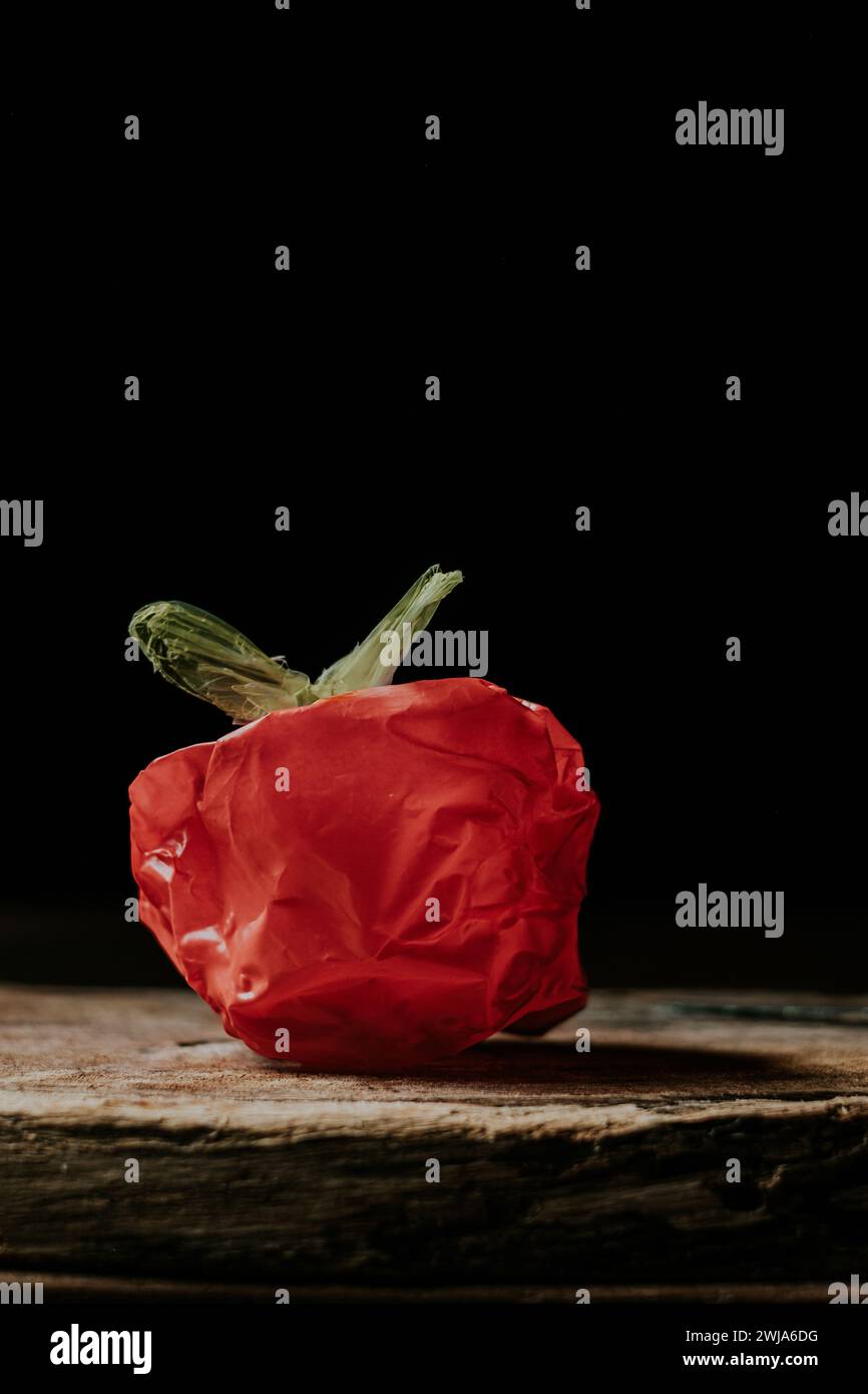 Eine rote Plastiktüte, die einem Gemüse oder einer Frucht ähnelt, auf einer Holzoberfläche vor schwarzem Hintergrund Stockfoto