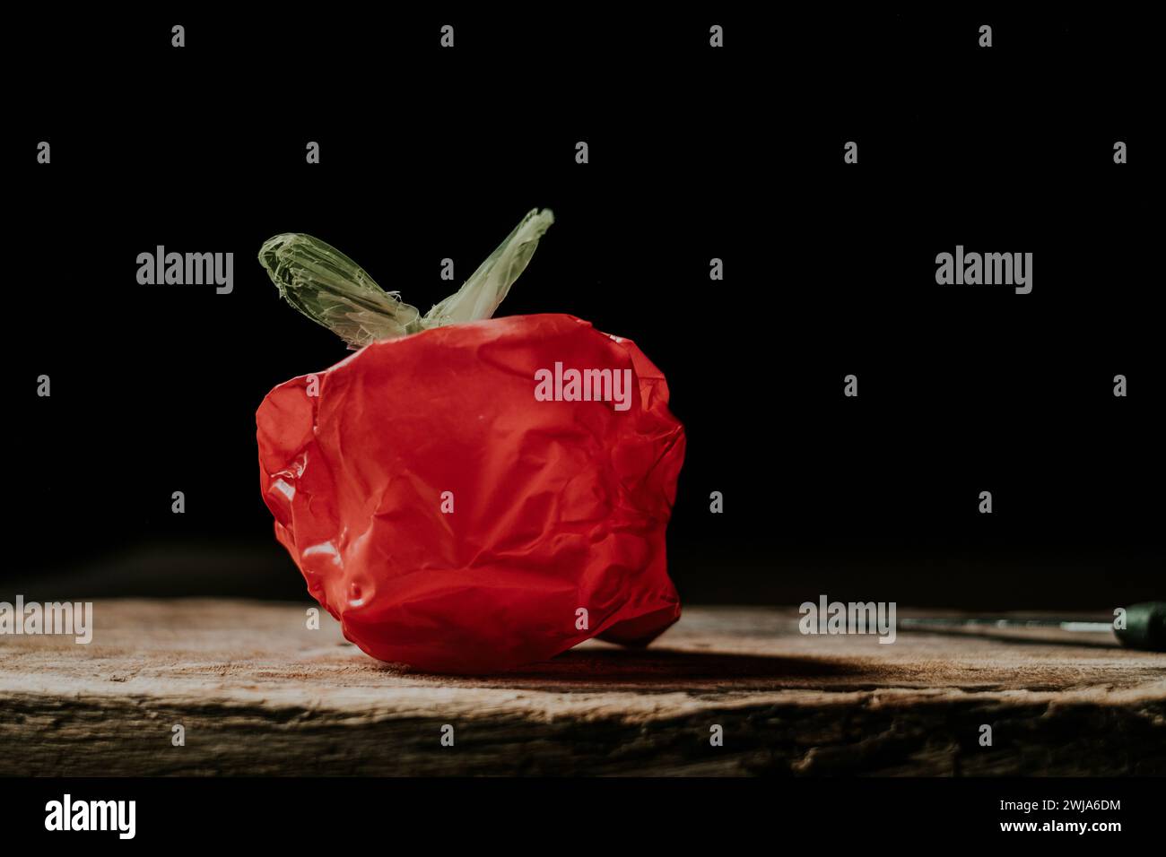 Eine rote Plastiktüte, die einem Gemüse oder einer Frucht ähnelt, auf einer Holzoberfläche vor schwarzem Hintergrund Stockfoto