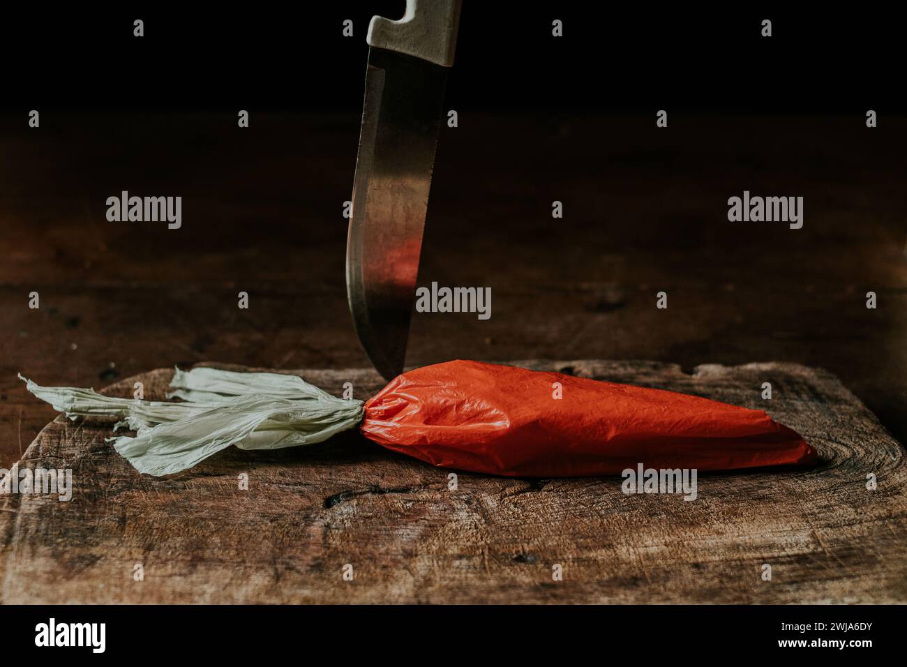 Eine Plastiktüte, die einer Karotte ähnelt und über ihr ein Küchenmesser auf einem abgenutzten Holzbrett platziert ist Stockfoto