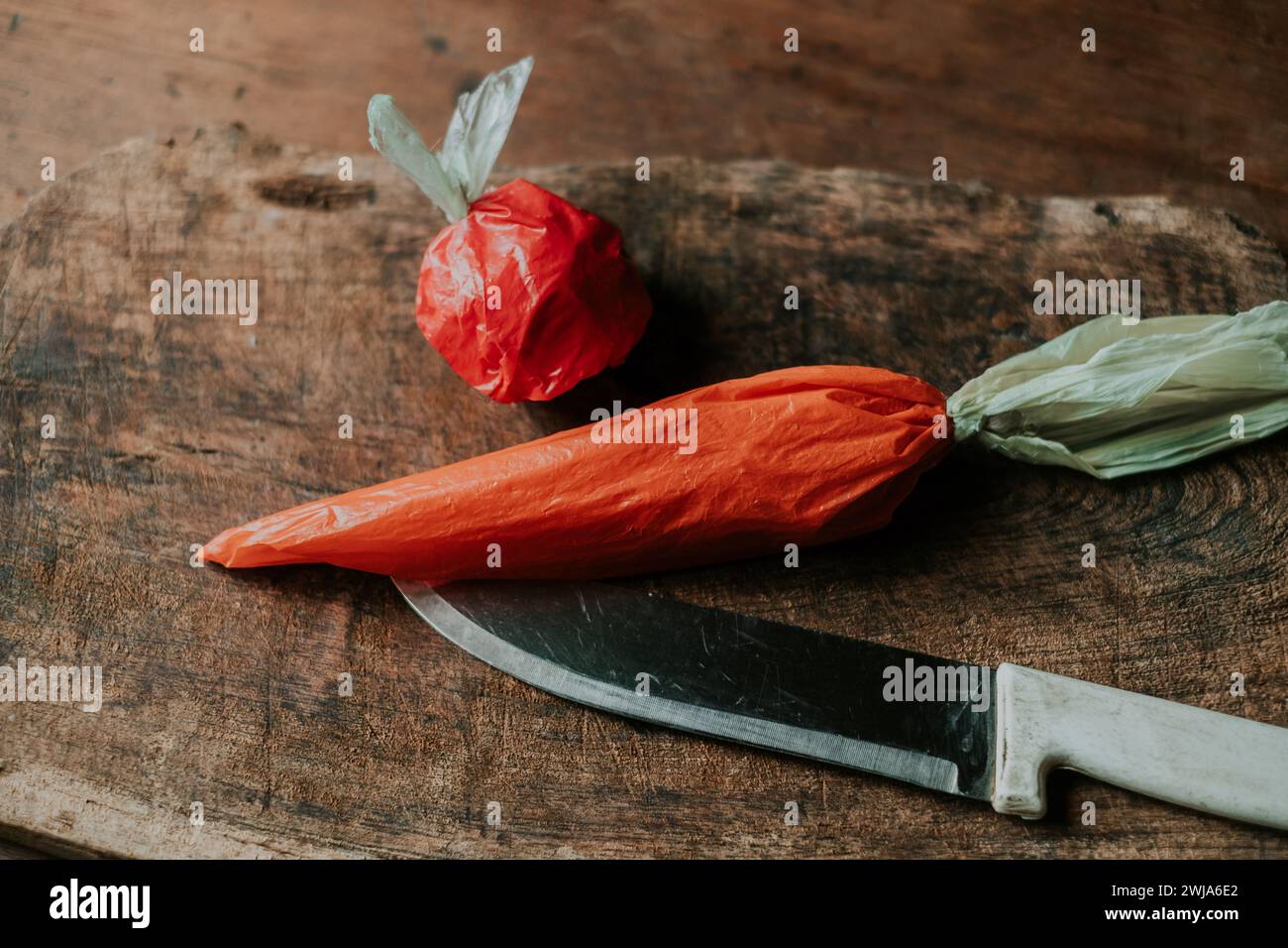 Ein roter Plastikbeutel in Form einer Tomate und ein orangener Beutel, der einer Karotte ähnelt, neben einem Messer auf einem Holzbrett Stockfoto