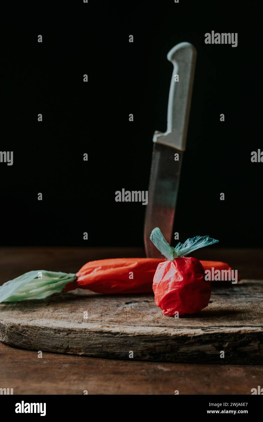 Plastiktüten, die an eine Tomate und Karotte erinnern, neben einem Küchenmesser auf einem rustikalen Holzschneidebrett Stockfoto