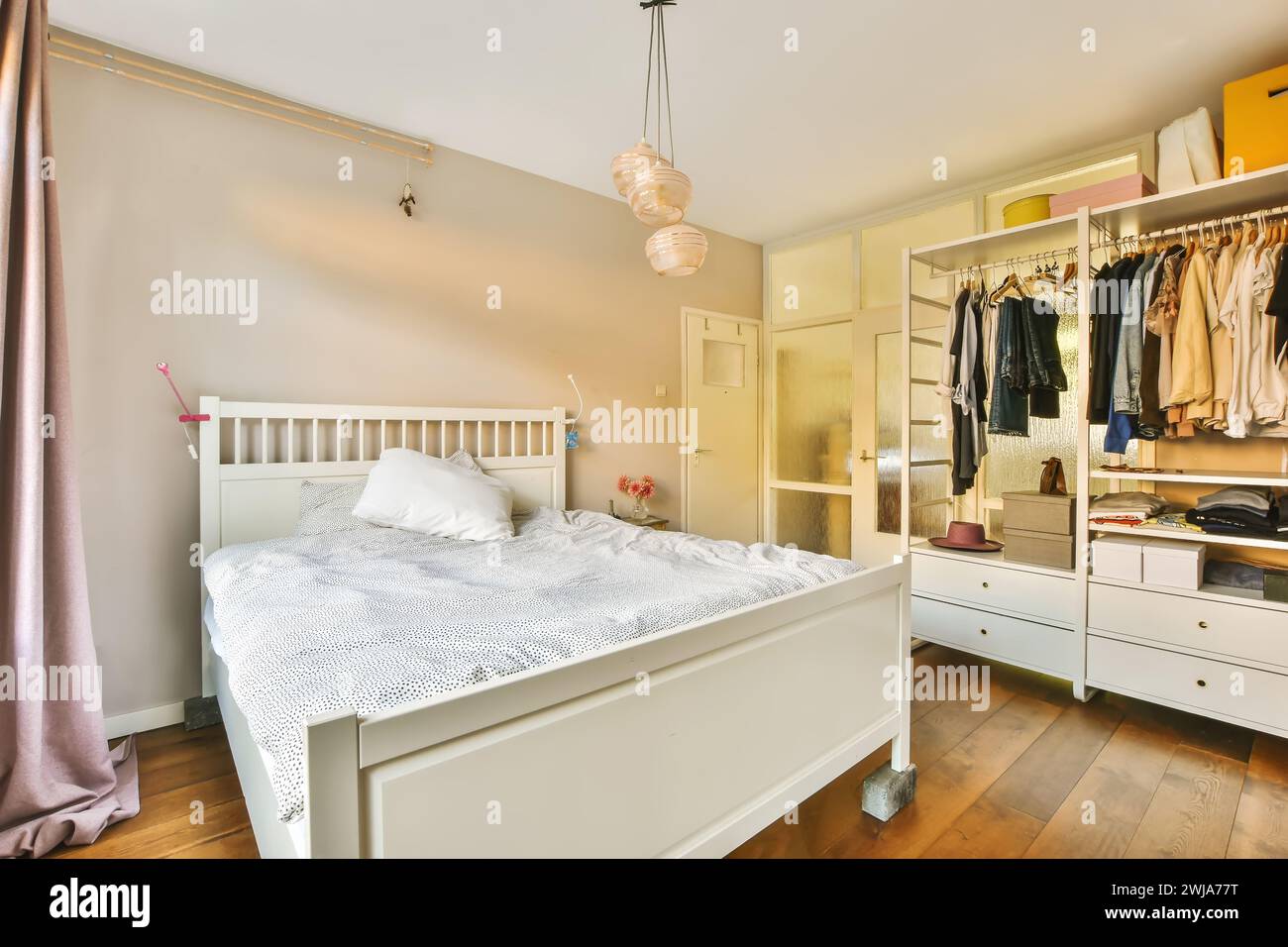 Ein komfortables und warm beleuchtetes Schlafzimmer mit weißem Bett, offenem Kleiderschrank mit Kleidung und bezaubernden Pendelleuchten Stockfoto