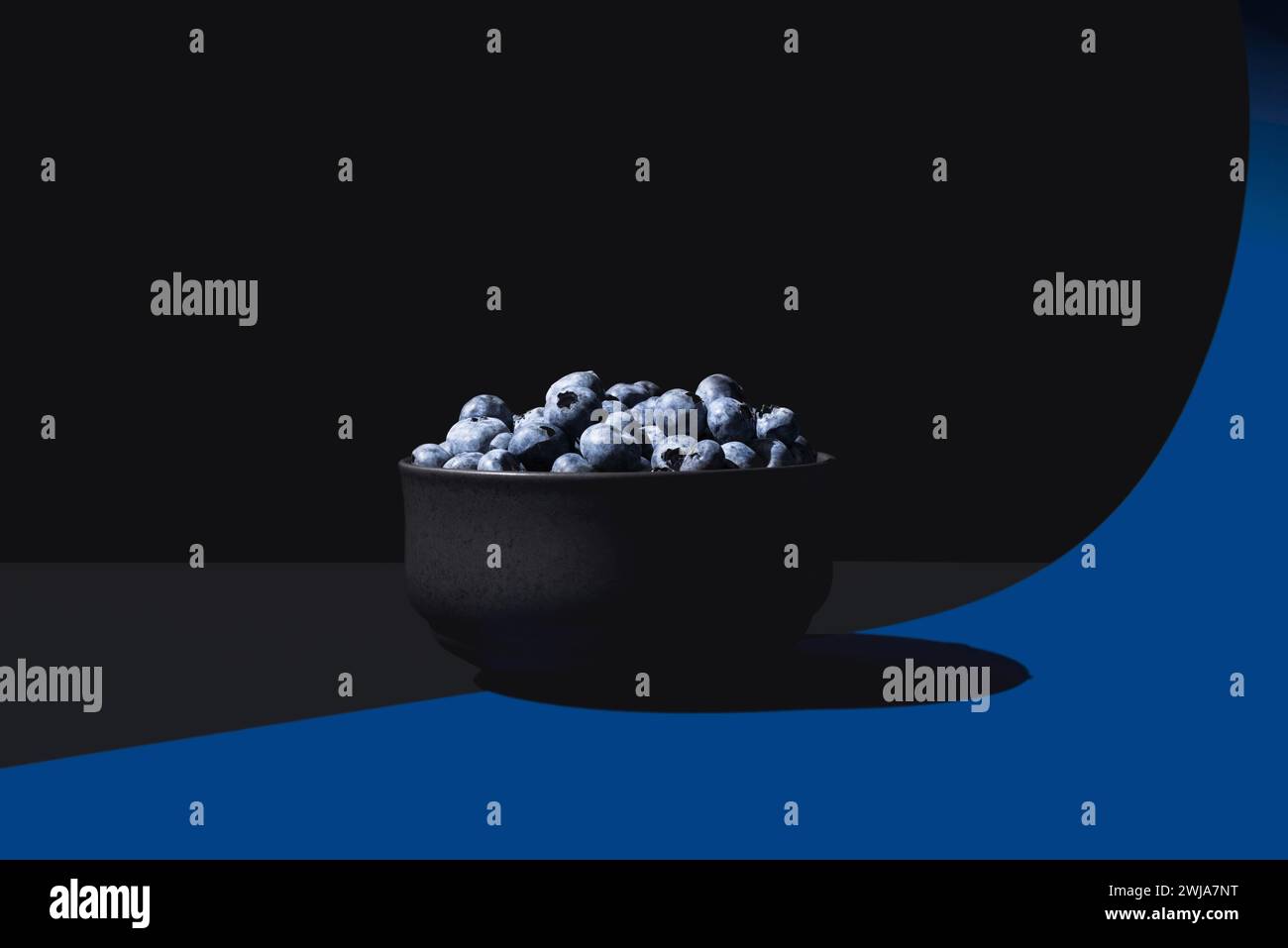Eine minimalistische Komposition mit einer schwarzen Schale Heidelbeeren auf einem geteilten blau-schwarzen Hintergrund mit dramatischer Beleuchtung Stockfoto
