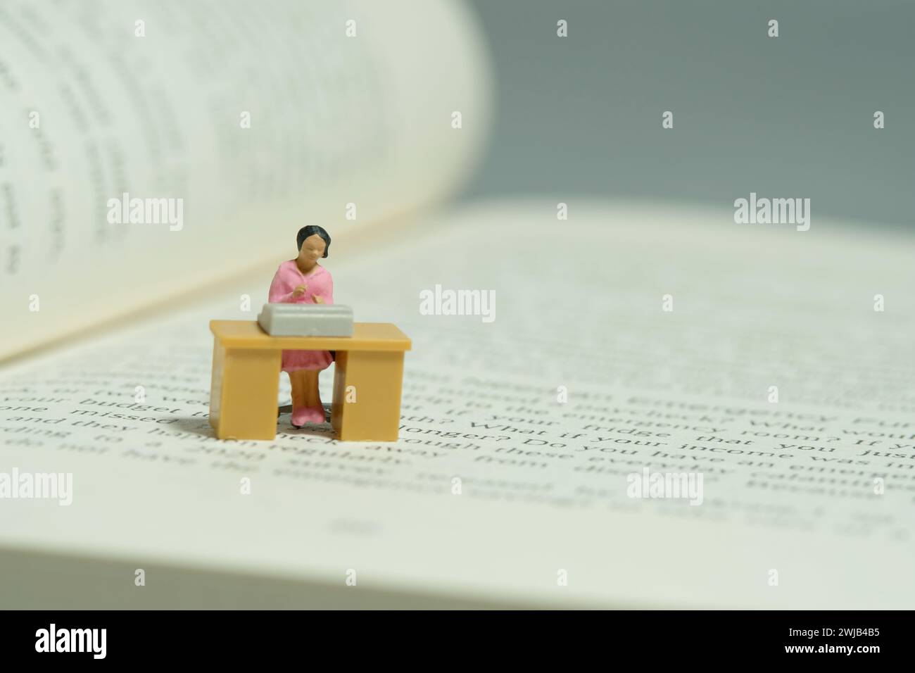 Miniatur-Kleinkinder-Spielfiguren-Fotografie. Literaturrecherche. Eine Frau sitzt auf dem Schreibtisch über dem geöffneten Buch. Bild Foto Stockfoto