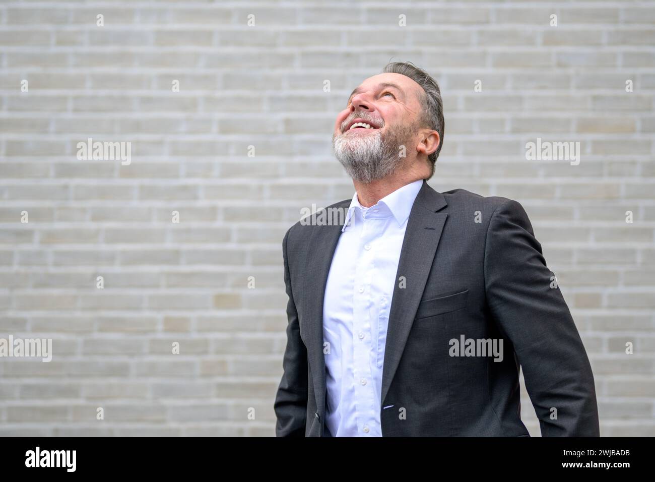 Attraktiver sechzigjähriger grauer Mann vor einer weißen Ziegelwand, der mit einem großen Lächeln aufblickt Stockfoto