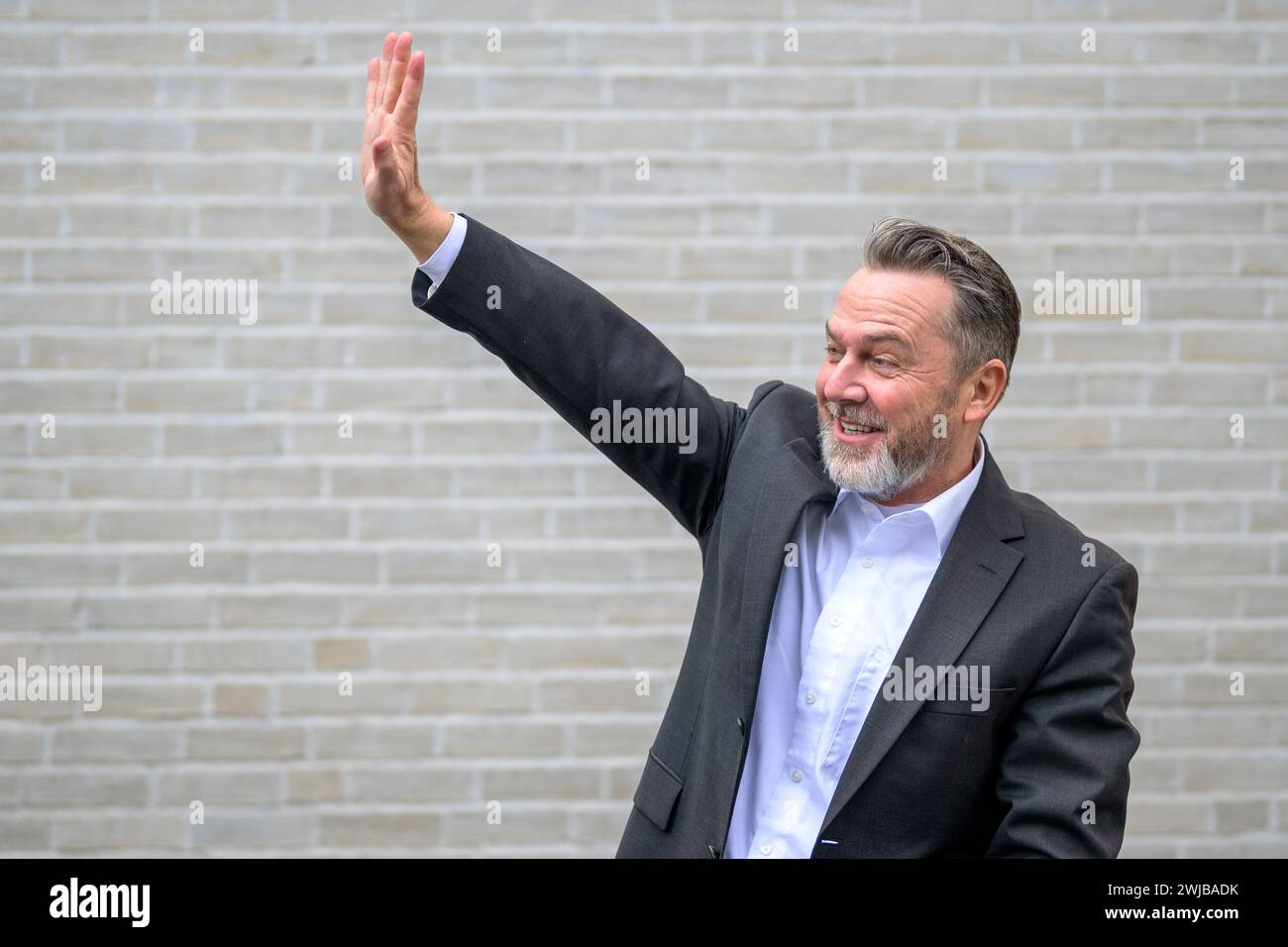 Attraktiver sechzigjähriger grauer Mann vor einer weißen Ziegelmauer, der jemandem in der Ferne zuwinkt Stockfoto