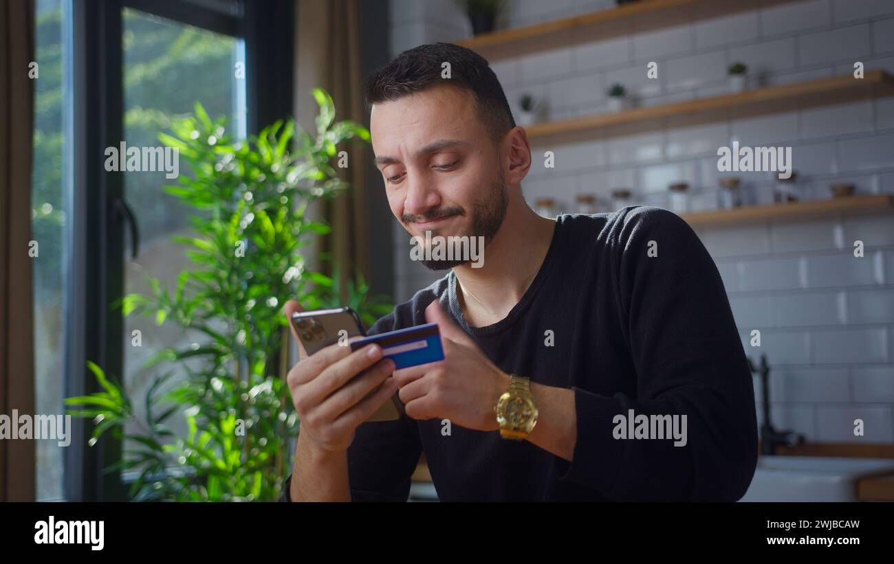 Erwachsener Mann mit Kreditkarte und Smartphone und aufgrund unzureichender Geldmittel Probleme bei der Online-Zahlung mit Kreditkarte, während er in Kitc sitzt Stockfoto