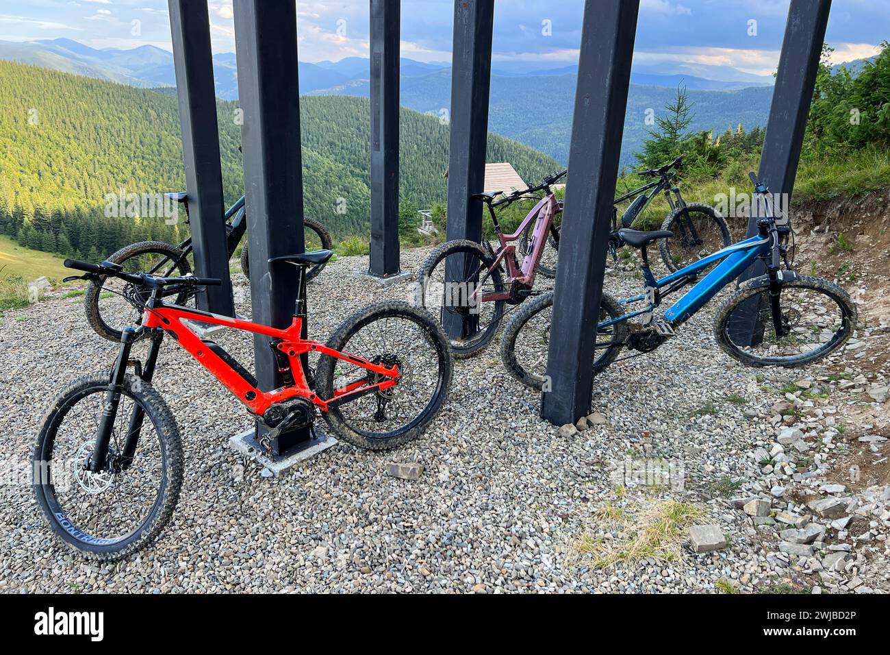Sammlung von Elektro-Mountainbikes, die auf einem kiesbedeckten Boden in den Bergen geparkt sind. MTB-Bikes befinden sich unter einer Struktur mit dicken schwarzen Säulen. Stockfoto