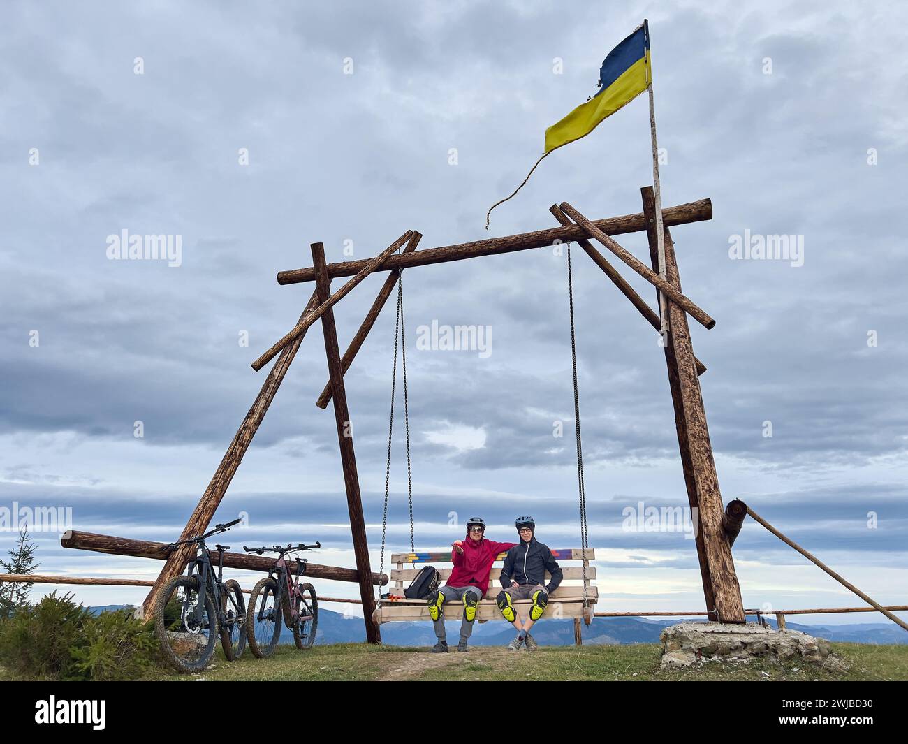 Zwei Männer, die vom Radfahren ausruhen, sitzen auf einer großen Holzschaukel draußen neben zwei elektrischen Mountainbikes. Karpaten, Ukraine. Stockfoto