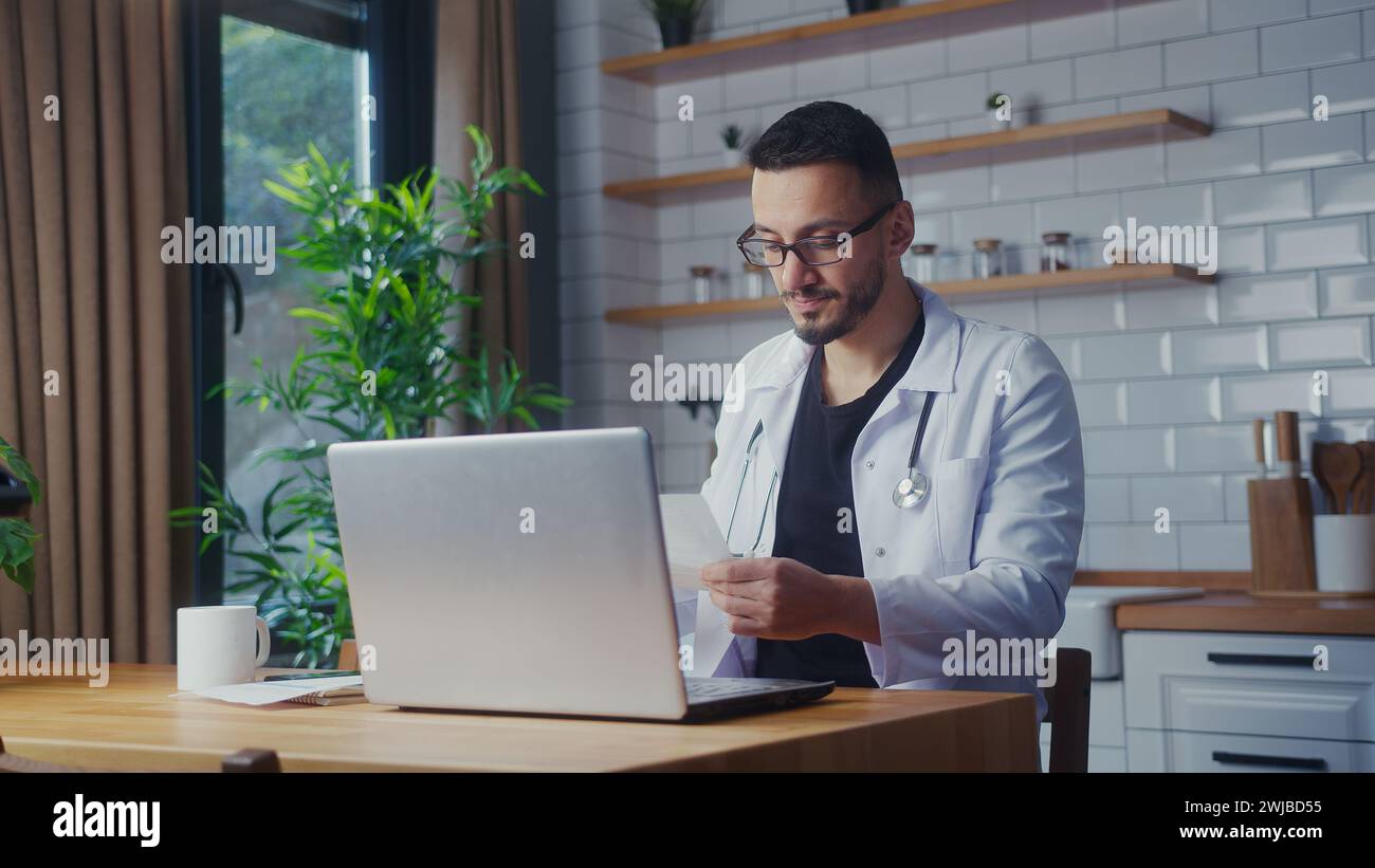 Erfahrener männlicher Arzt mit Brille mit weißem Mantel mit Laptop-Computer, der Patientenberichte im Heimbüro studiert. Männlicher Praktizierender, der Papierkram macht Stockfoto