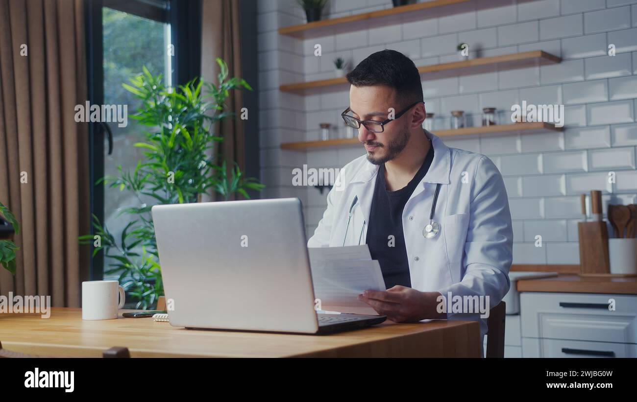 Ärztin mit Brille im weißen Mantel mit Laptop-Computer, die Patientenberichte im Heimbüro untersucht. Männlicher Praktizierender, der Papierkram macht Stockfoto