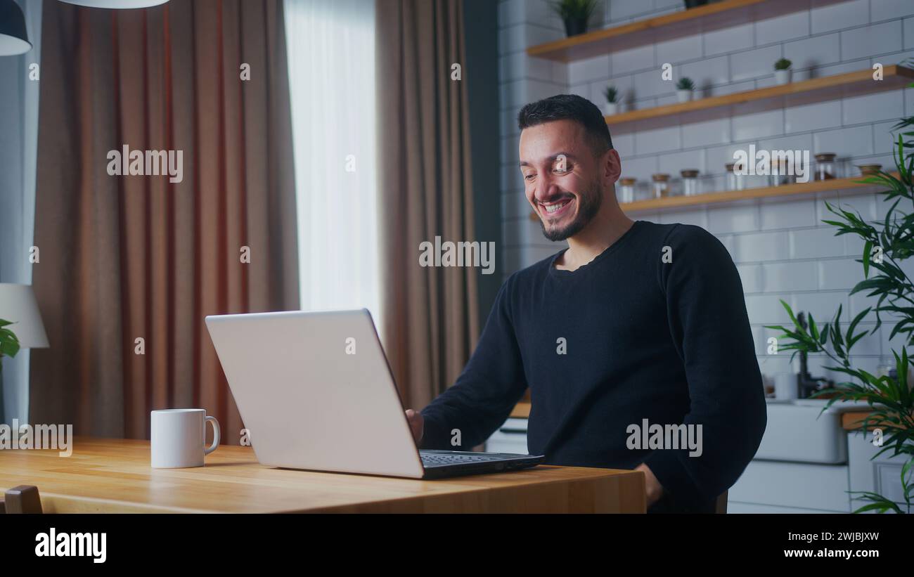 Mann, der in der Küche sitzt und E-Mails liest, Nachricht lesen, gute Nachrichten erhalten Lotterie auf Laptop gewinnen große Chance feiern fühlt sich glücklich Stockfoto