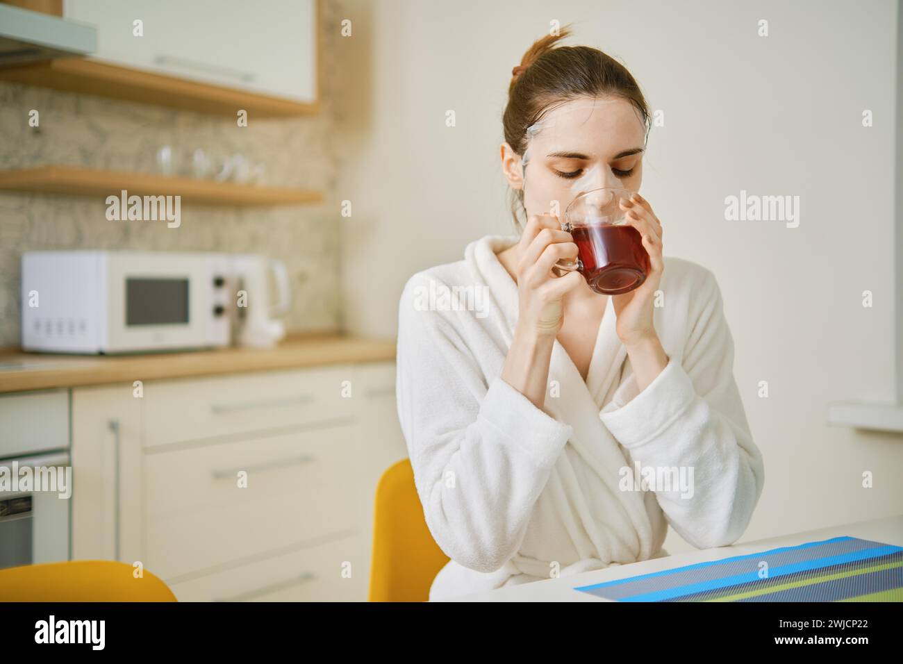 Eine Frau mit einer kosmetischen Anti-Aging-Maske im Gesicht sitzt in der Küche und trinkt Tee Stockfoto