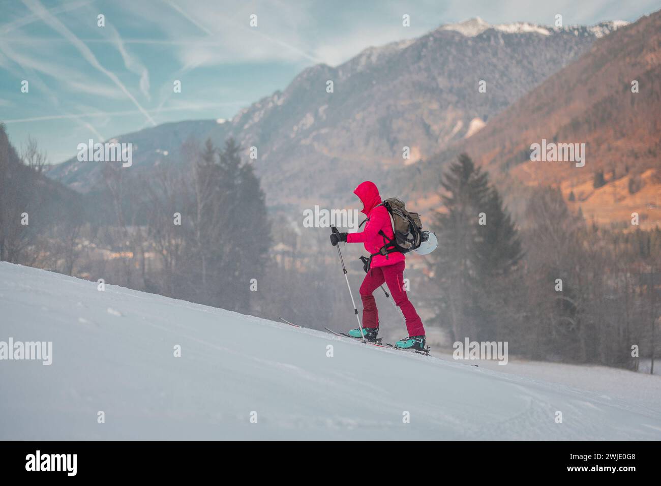 Skitourenfrau in rosa Kleidung steigt langsam auf einer verschneiten Skipiste mit majestätischen Bergen im Hintergrund an. Seitenansicht eines Skibergs Stockfoto