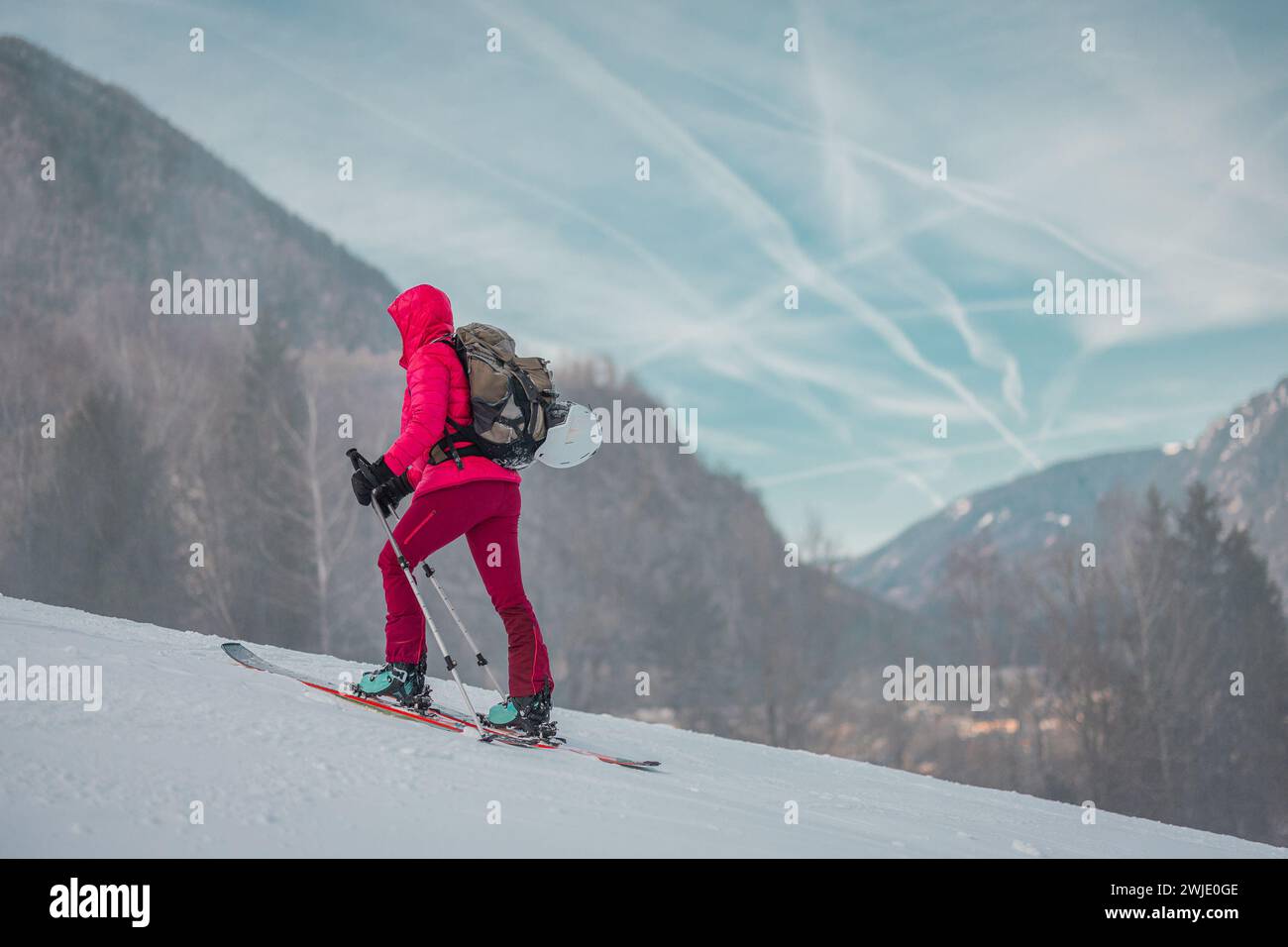Skitourenfrau in rosa Kleidung steigt langsam auf einer verschneiten Skipiste mit majestätischen Bergen im Hintergrund an. Seitenansicht eines Skibergs Stockfoto