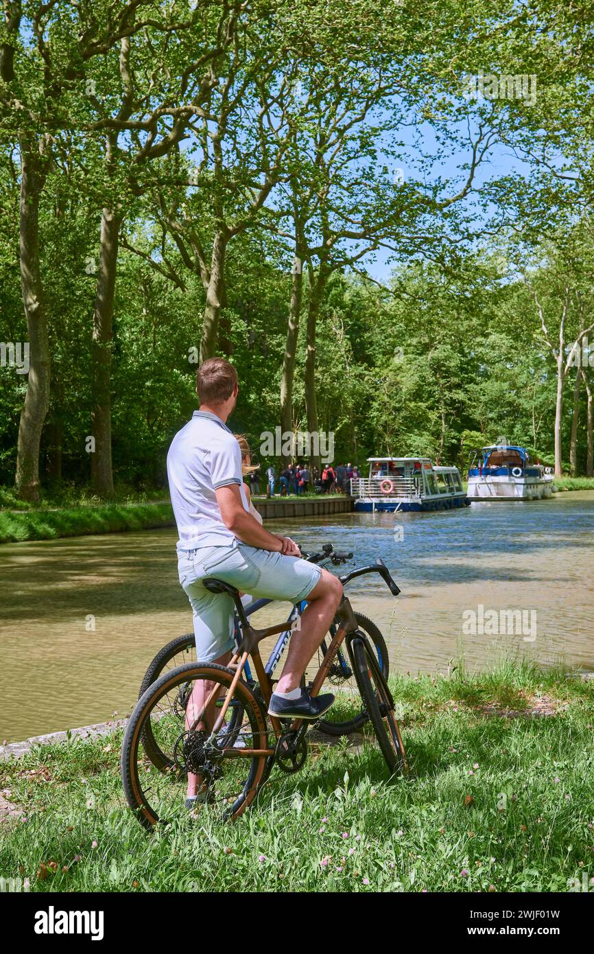 Ein Paar auf einer Fahrradtour entlang des Kanals, mit einem Lastkahn und Schleusenwärter, das in das Restaurant „La Bonne Planque“ umgewandelt wurde. Der Canal du Midi ist Registere Stockfoto