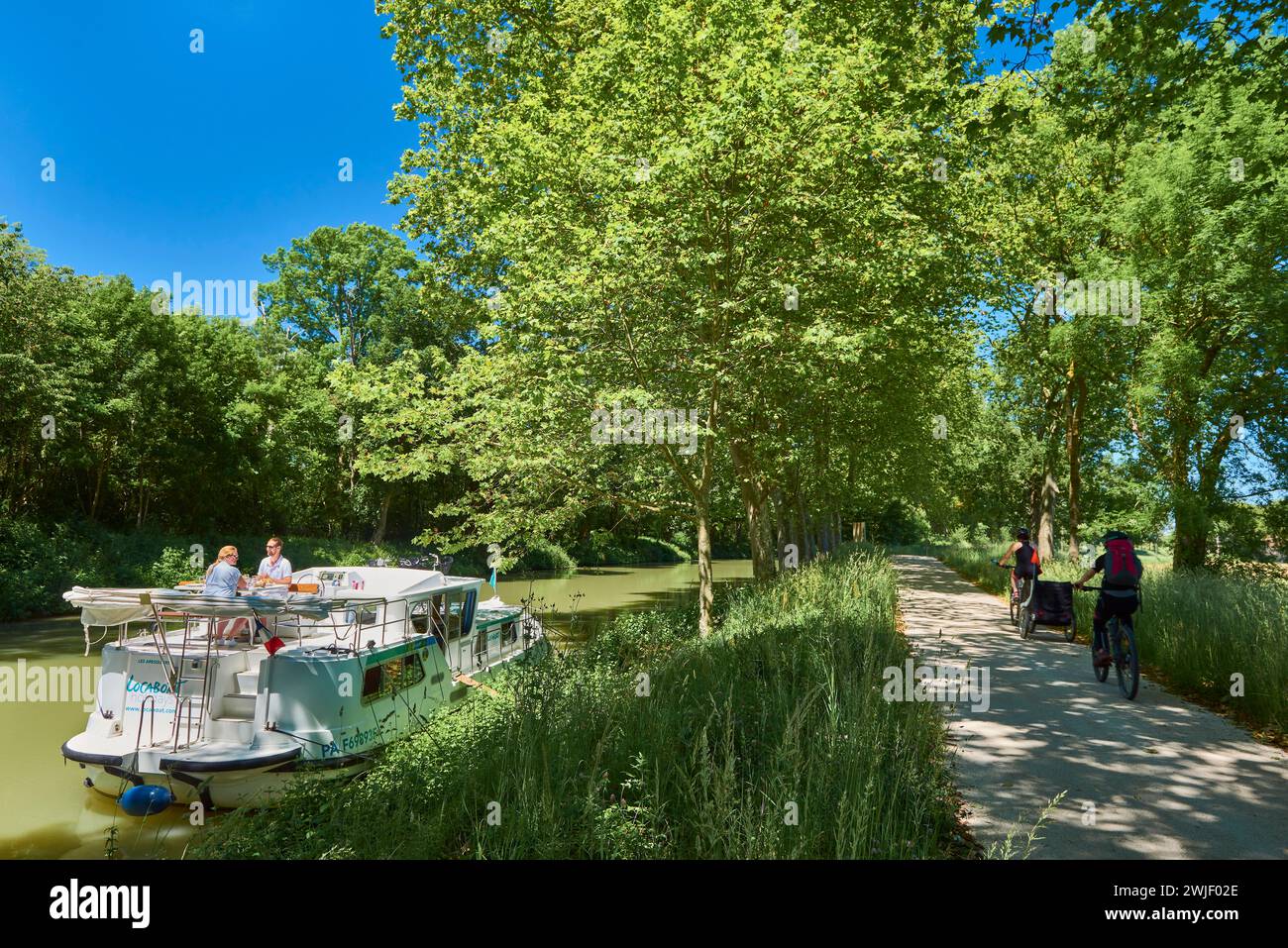 Ein Paar auf einem Mietkahn am Canal du Midi. Essen an Bord nach dem Anlegen am Ufer im Schatten der Platanen. Touristen, die Fahrrad fahren Stockfoto