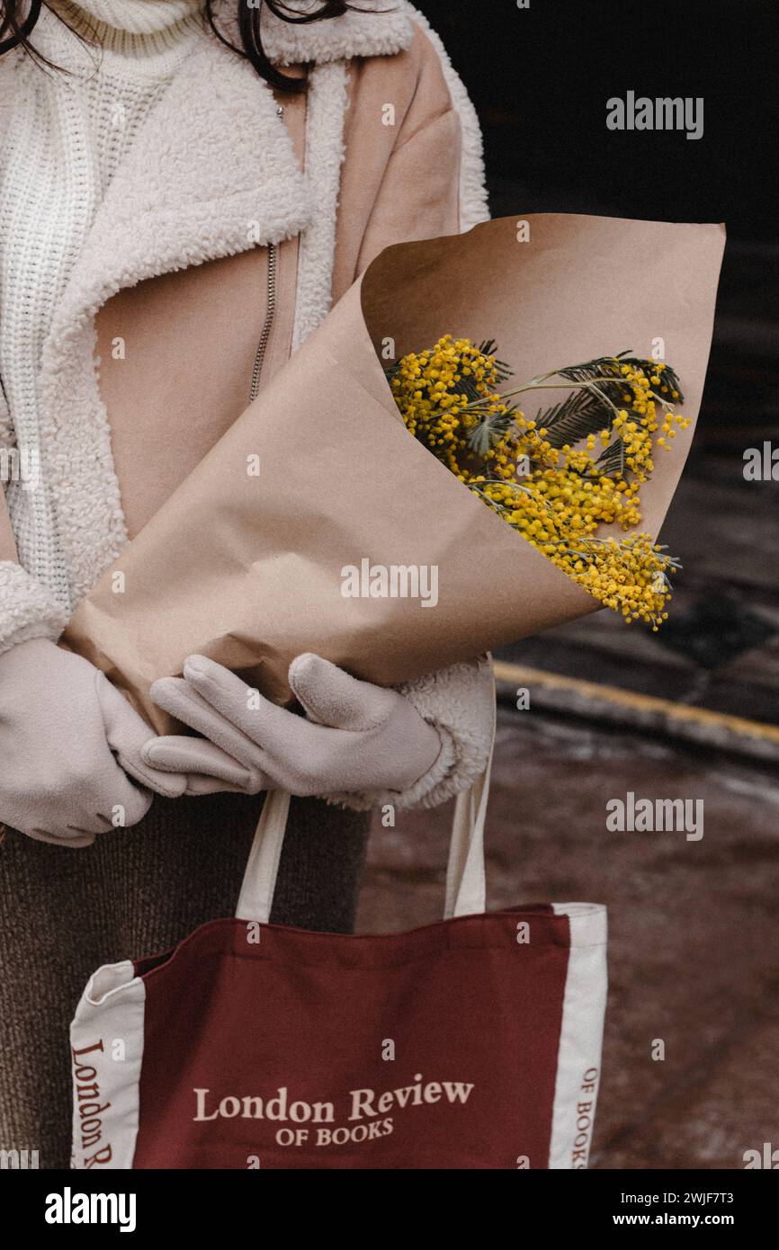 Eine Frau in einer beigen warmen Jacke und Pelzhandschuhen, die einen Strauß frischer Mimosa-Blüten hält. Stylischer Winter-Look Stockfoto
