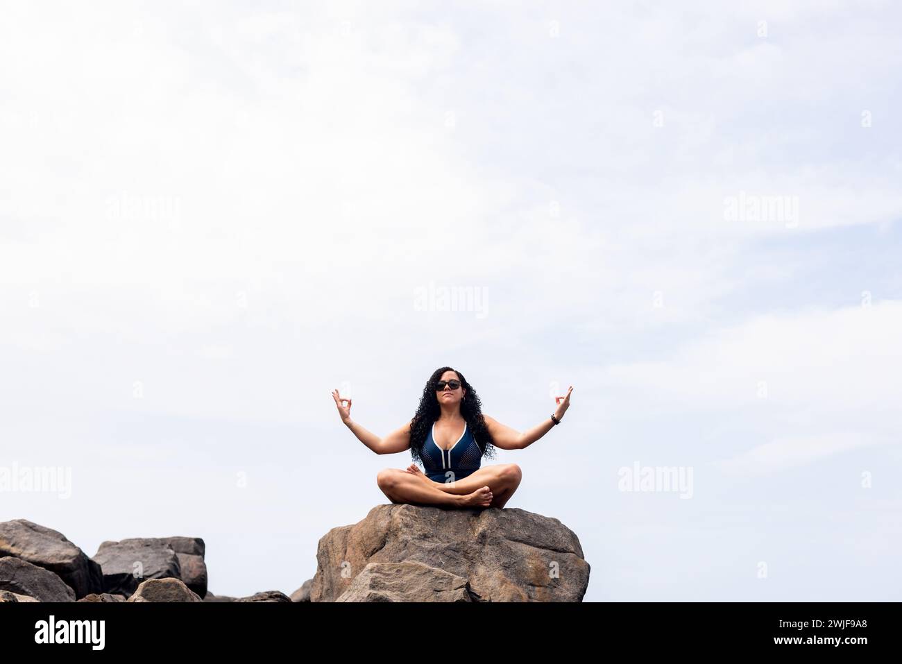 Salvador, bahia, Brasilien - 06. Januar 2024: Schöne, lockige Frau, die auf dem Felsen eines Strandes sitzt und Yoga-Übungen macht. Glückliche Person auf Reisen. Stockfoto