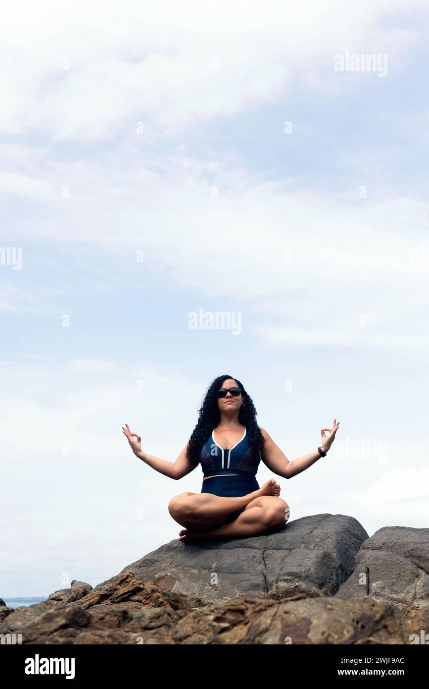 Salvador, bahia, Brasilien - 06. Januar 2024: Schöne, lockige Frau, die auf dem Felsen eines Strandes sitzt und Yoga-Übungen macht. Glückliche Person auf Reisen. Stockfoto