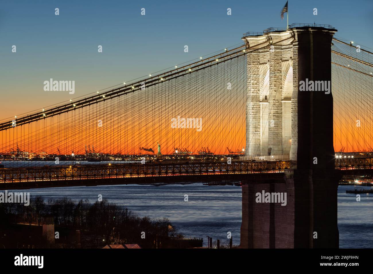 Beleuchtete Brooklyn Bridge bei Sonnenuntergang und die Freiheitsstatue in der Ferne. Nahaufnahme historischer Sehenswürdigkeiten von New York City Stockfoto