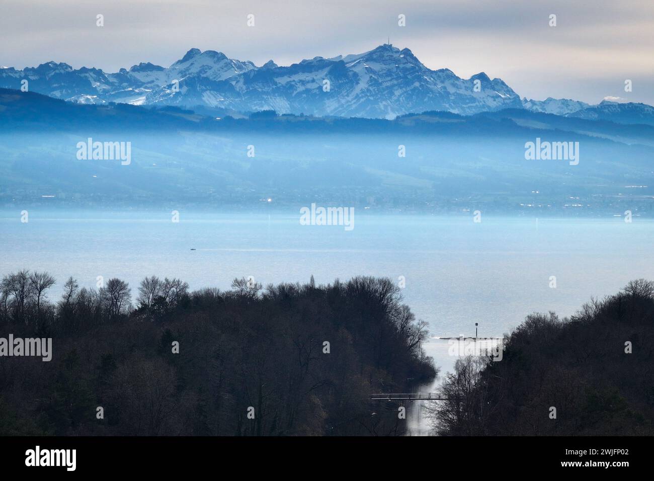 Langenargen, Deutschland. Februar 2024. Der Säntis mit seinem Mast auf dem Gipfel erhebt sich über den Dunst hinter dem Bodensee. Die Schweiz ist im Hintergrund zu sehen, im Vordergrund der Fluss Argen, der in den Bodensee mündet. (Luftaufnahme mit einer Drohne) Credit: Felix Kästle/dpa/Alamy Live News Stockfoto