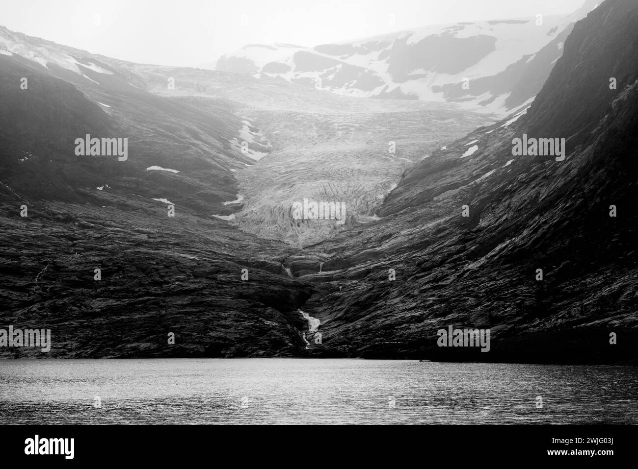 In diesem monochromen Bild entfaltet sich das strukturierte Eis des Svartisen-Gletschers in einem dunklen See, der von Norwegens dramatischem Berggebiet (schwarz und weiß) umhüllt wird Stockfoto