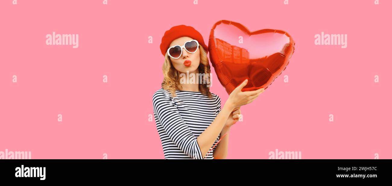 Süßes Porträt der schönen glücklichen jungen Frau mit rotem Herzballon bläst Kuss im französischen Baskenmütze auf rosa Studiohintergrund Stockfoto